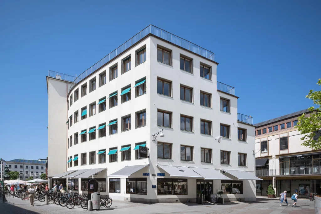 En vit fyrkantig byggnad med flera våningar och gröna markiser över fönstren.