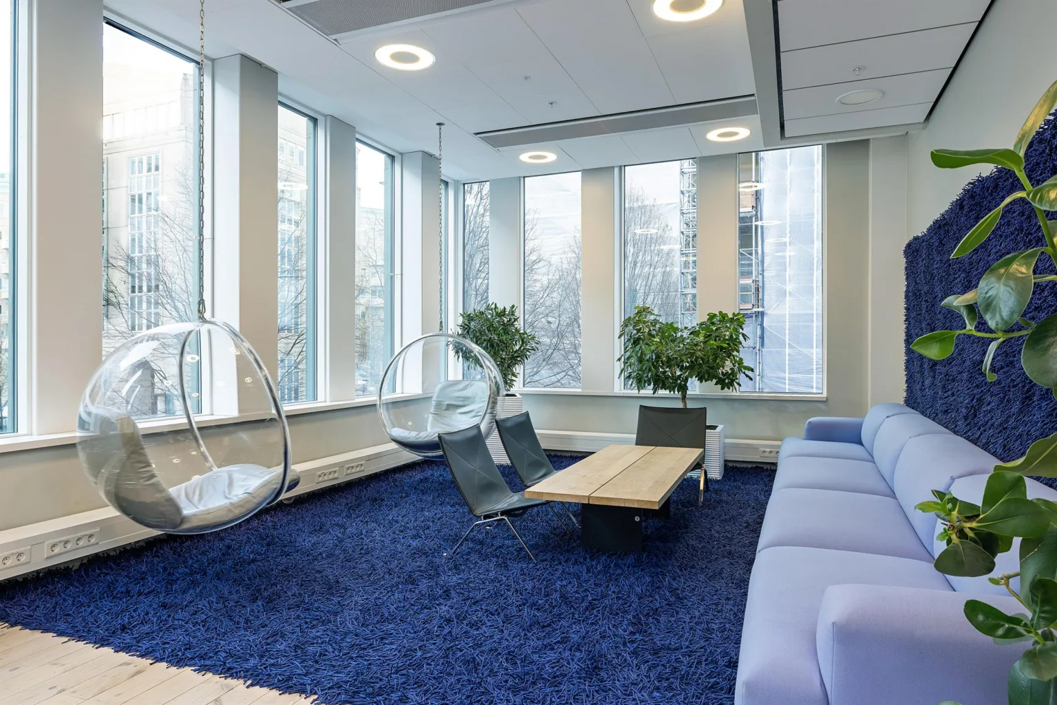 En lounge med en blå matta, en soffa, två stolar och hängande stolar bredvid stora fönster och gröna växter.