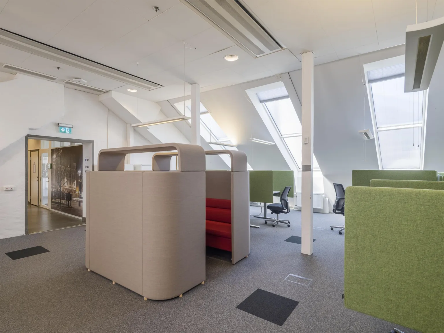 Öppet kontorslandskap med arbetsstationer, gröna avskärmningsväggar och takfönster som släpper in dagsljus.