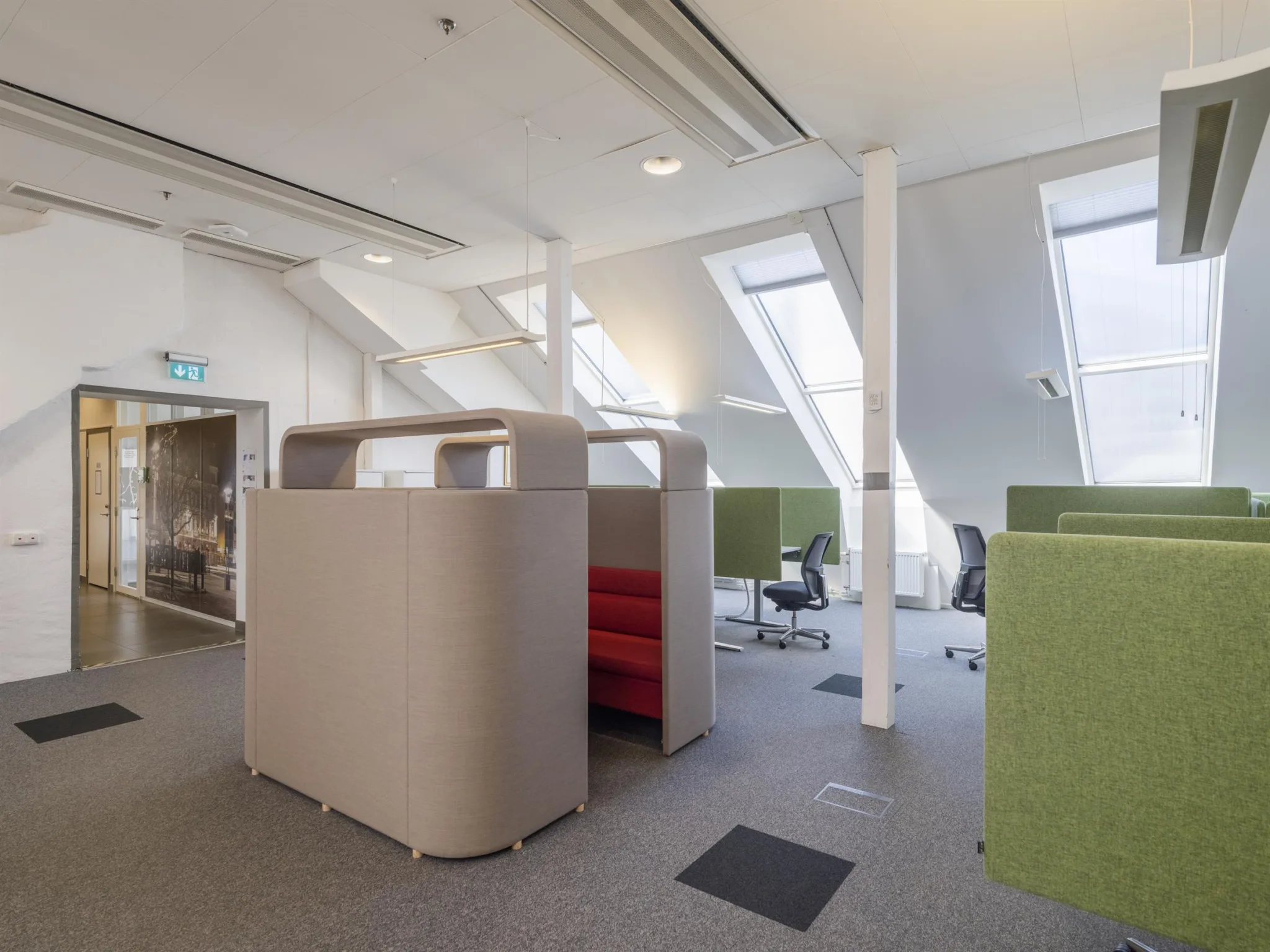 Öppet kontorslandskap med arbetsstationer, gröna avskärmningsväggar och takfönster som släpper in dagsljus.