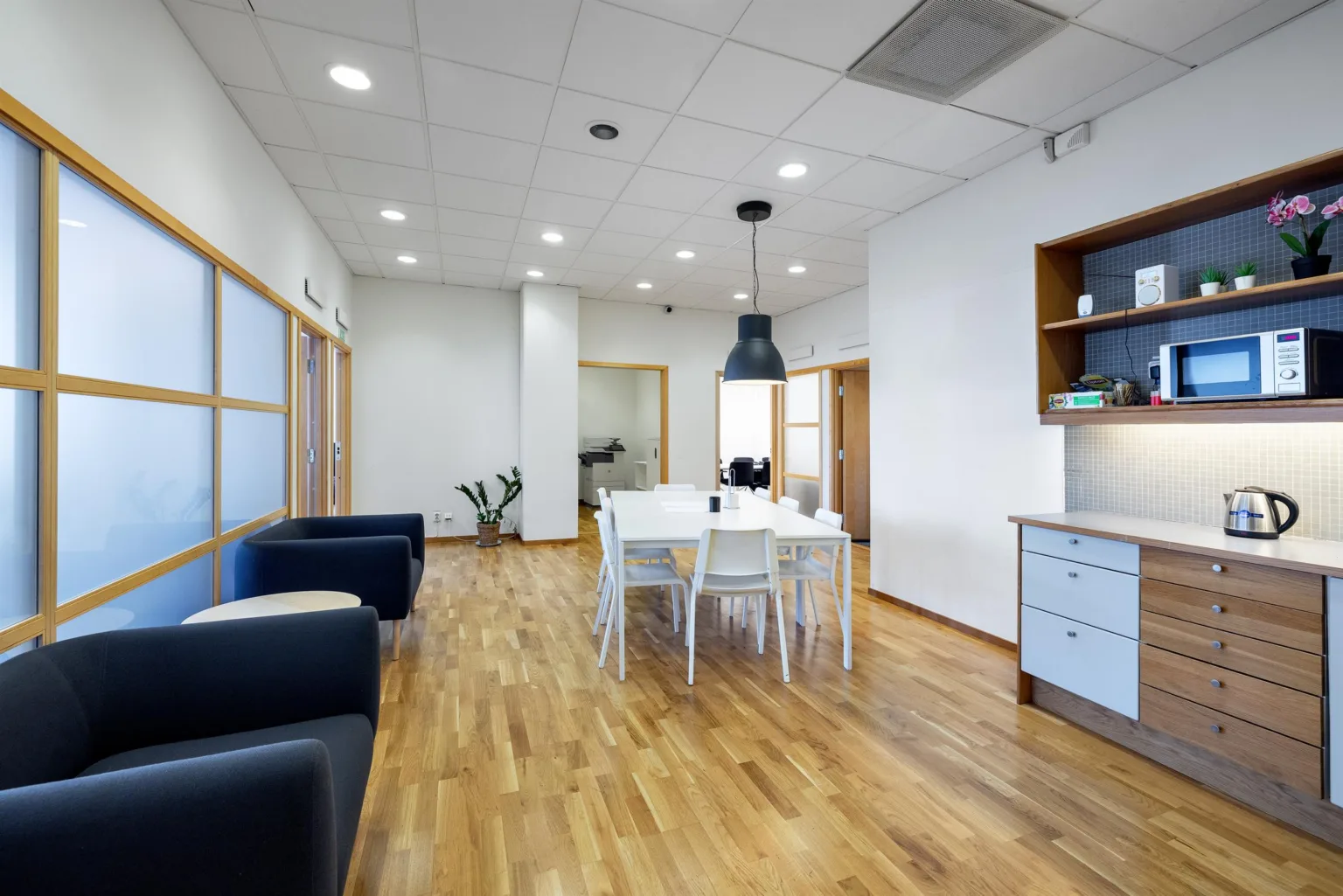 Ett ljust kontorsutrymme med trägolv, en matgrupp med vita stolar och en svart lampa ovanför samt soffor längs väggen.
