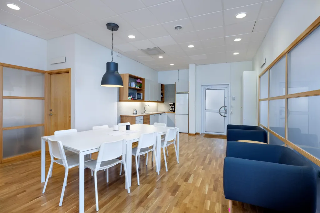 Ett köksutrymme med en matgrupp, vita stolar, en svart lampa ovanför och blåa soffor vid sidan.