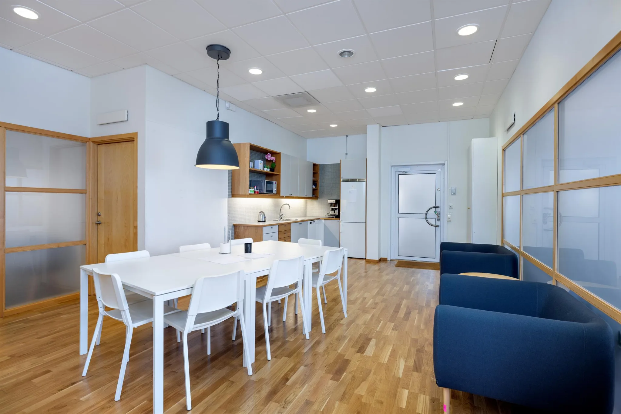 Ett köksutrymme med en matgrupp, vita stolar, en svart lampa ovanför och blåa soffor vid sidan.