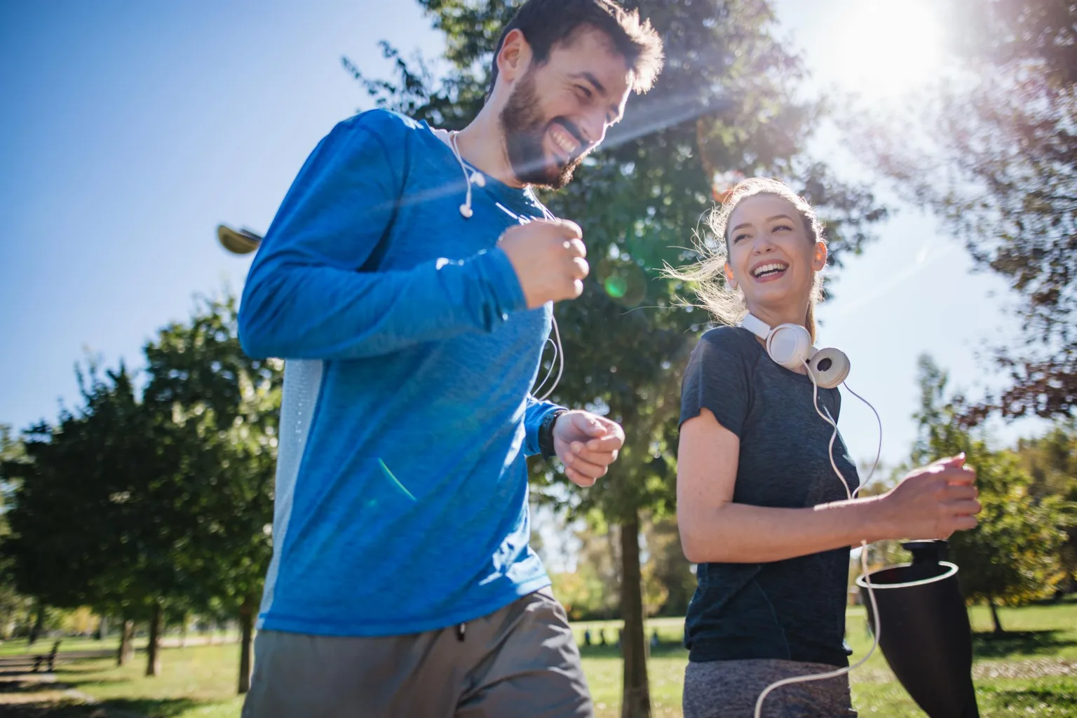 Två personer joggar utomhus i en solig park med träd och klarblå himmel.