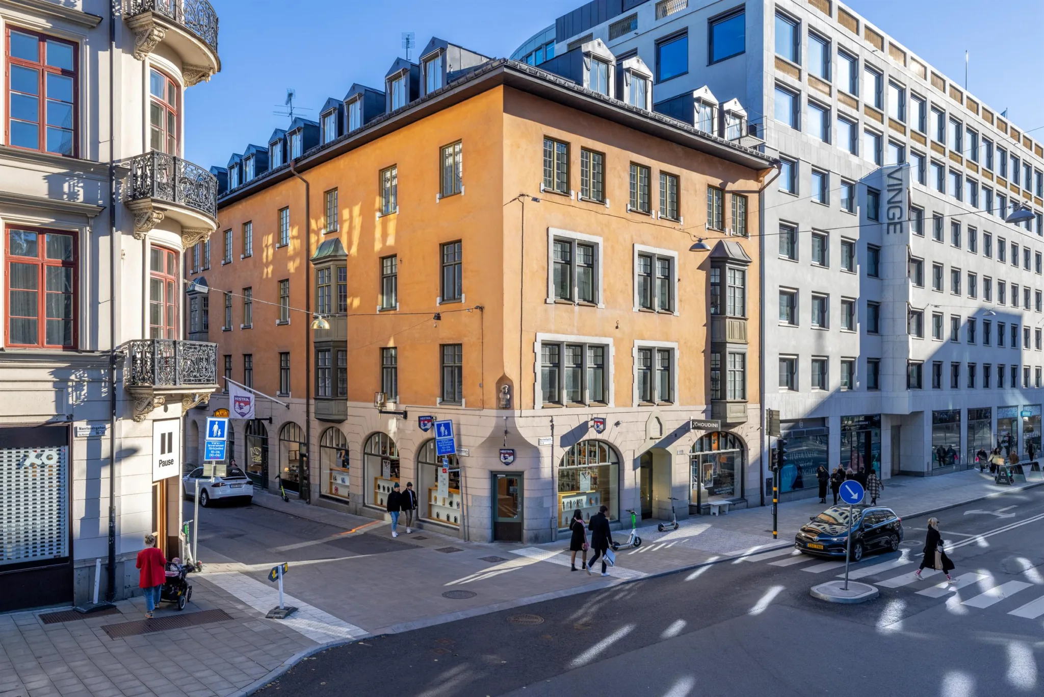 1. En orange byggnad med stora fönster belägen vid en korsning, omgiven av moderna byggnader och trottoarer med fotgängare.