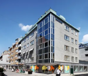 En hörnbyggnad med glasfasad och flera våningar, omgiven av mindre byggnader och människor som promenerar.