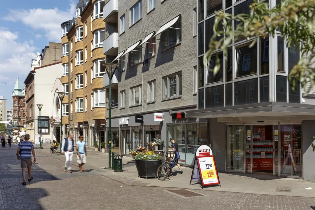 En shoppinggata med butiker, skyltar och människor som promenerar; färgglada blomkrukor står längs trottoaren.