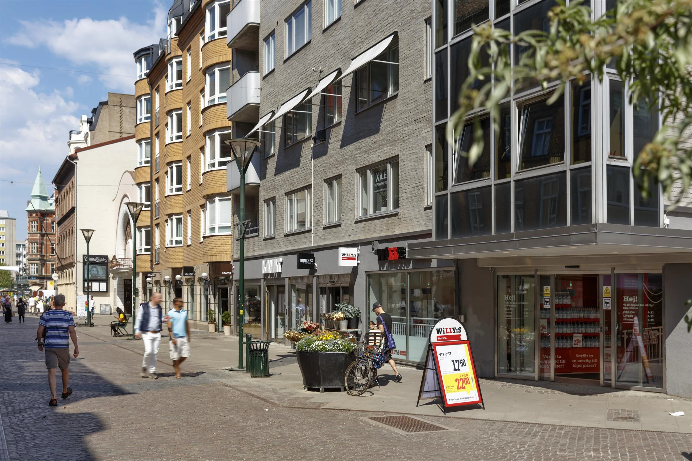En shoppinggata med butiker, skyltar och människor som promenerar; färgglada blomkrukor står längs trottoaren.