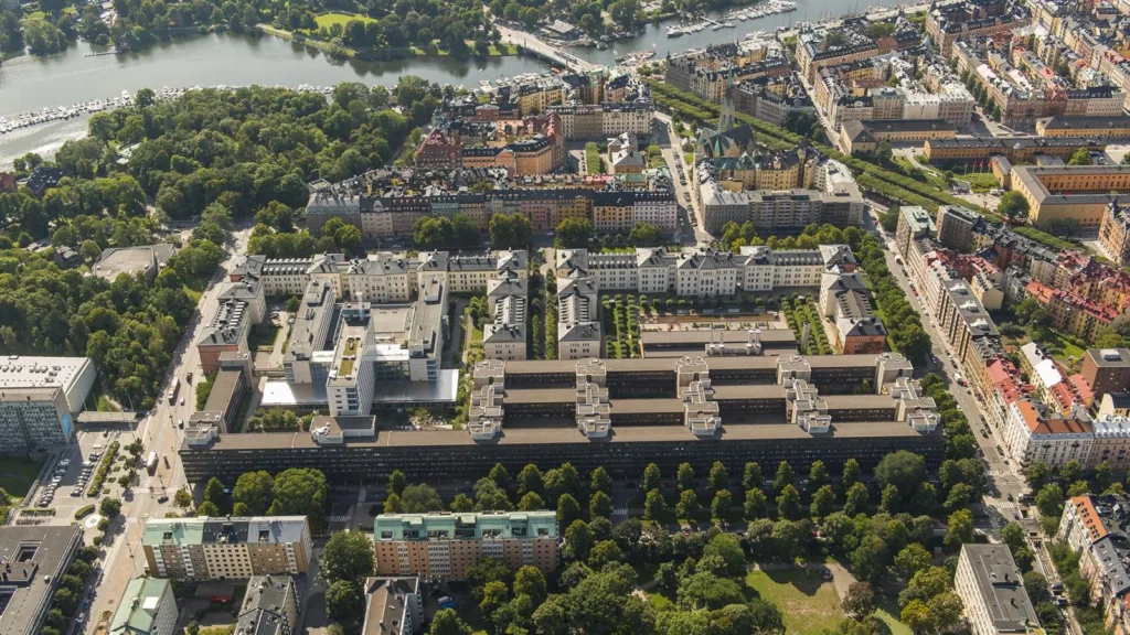 Flygfoto över ett område med symmetriska byggnader, grönområden, och närliggande vatten.