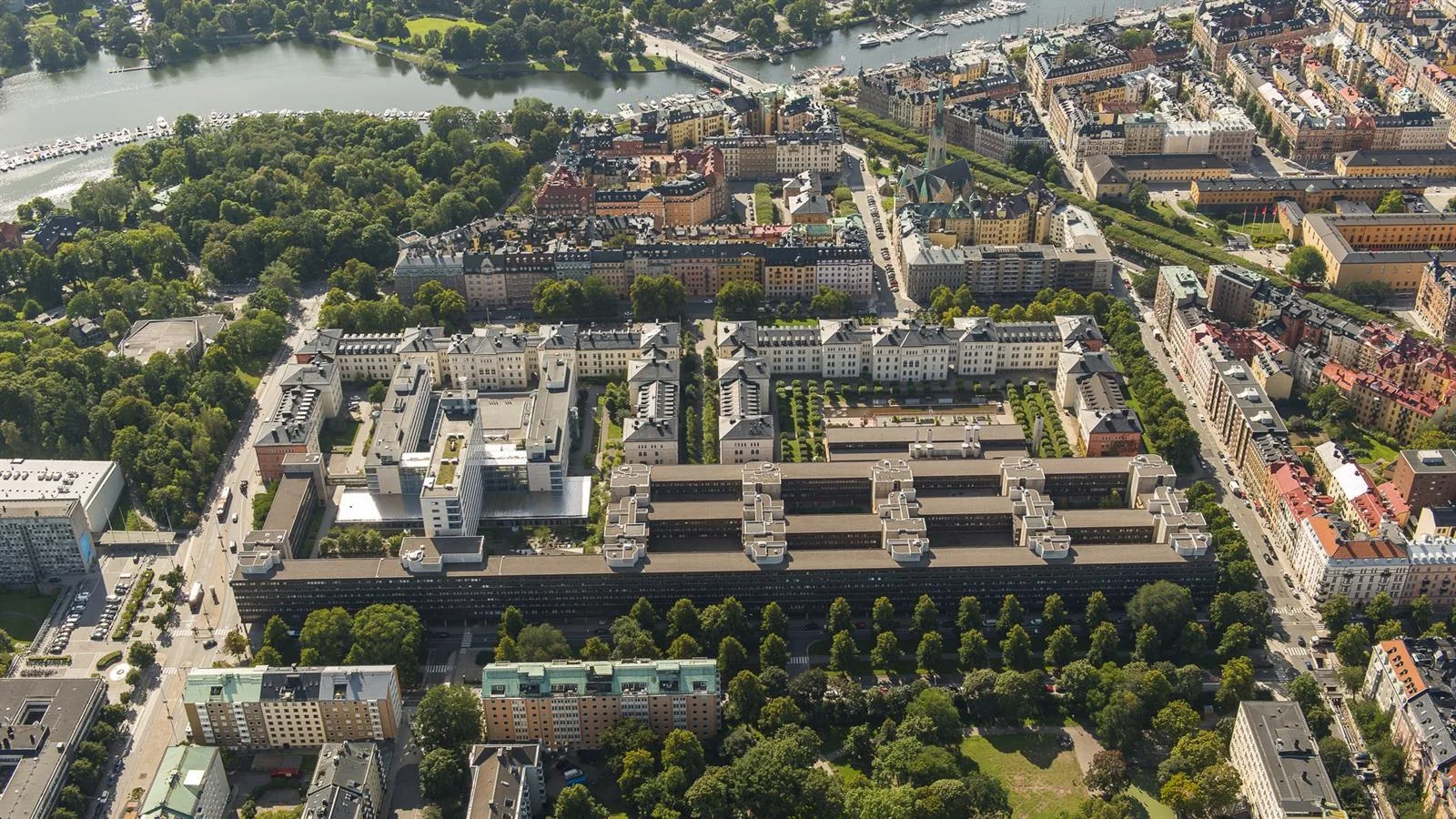 Flygfoto över ett område med symmetriska byggnader, grönområden, och närliggande vatten.