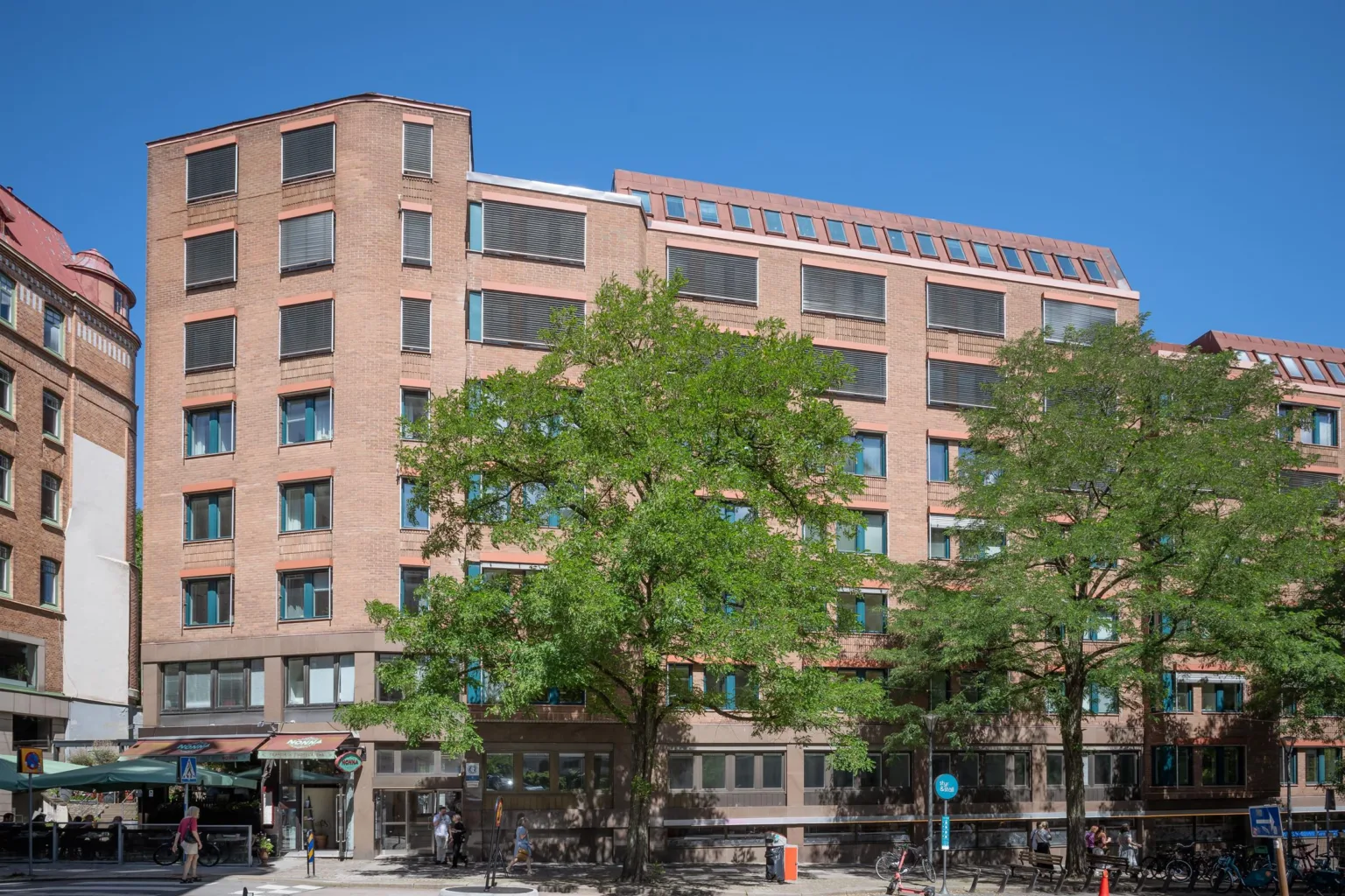 En brun tegelbyggnad med flera våningar, stora fönster och grönskande träd framför.
