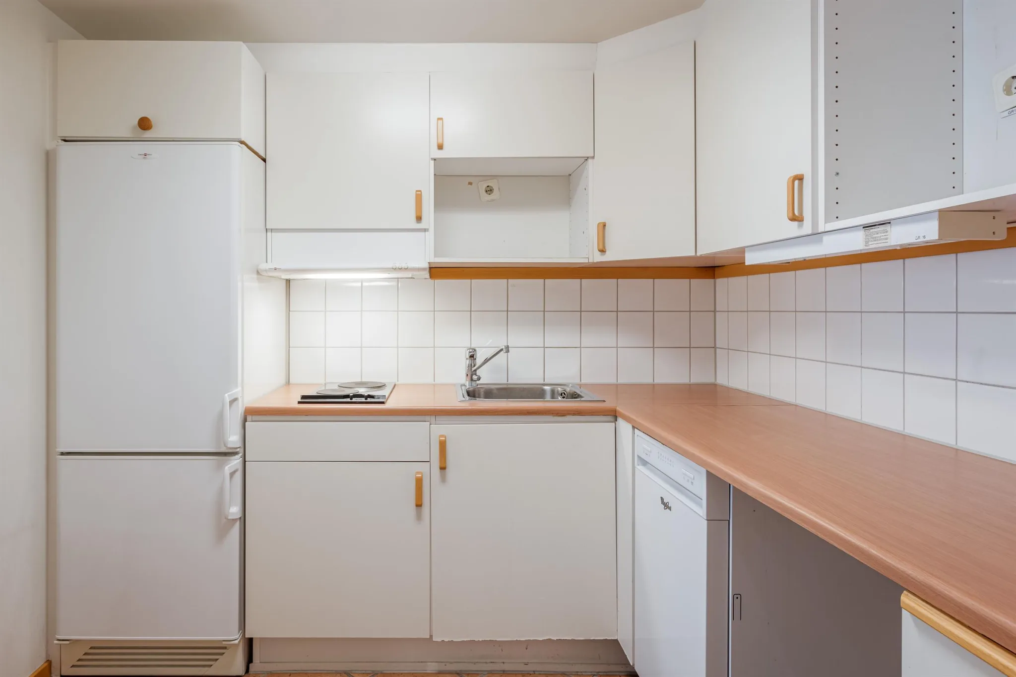 Ett vitt kök med skåp, en kyl, diskho och en bänkskiva i träfärg.