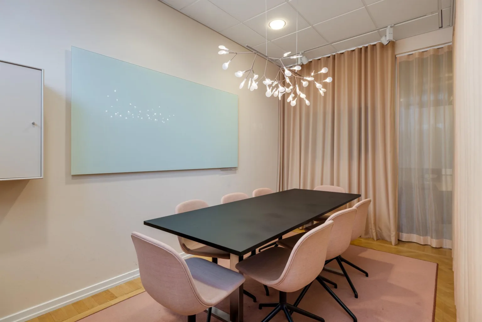 Ett mötesrum med ett svart bord, rosa stolar och en modern ljuskrona.