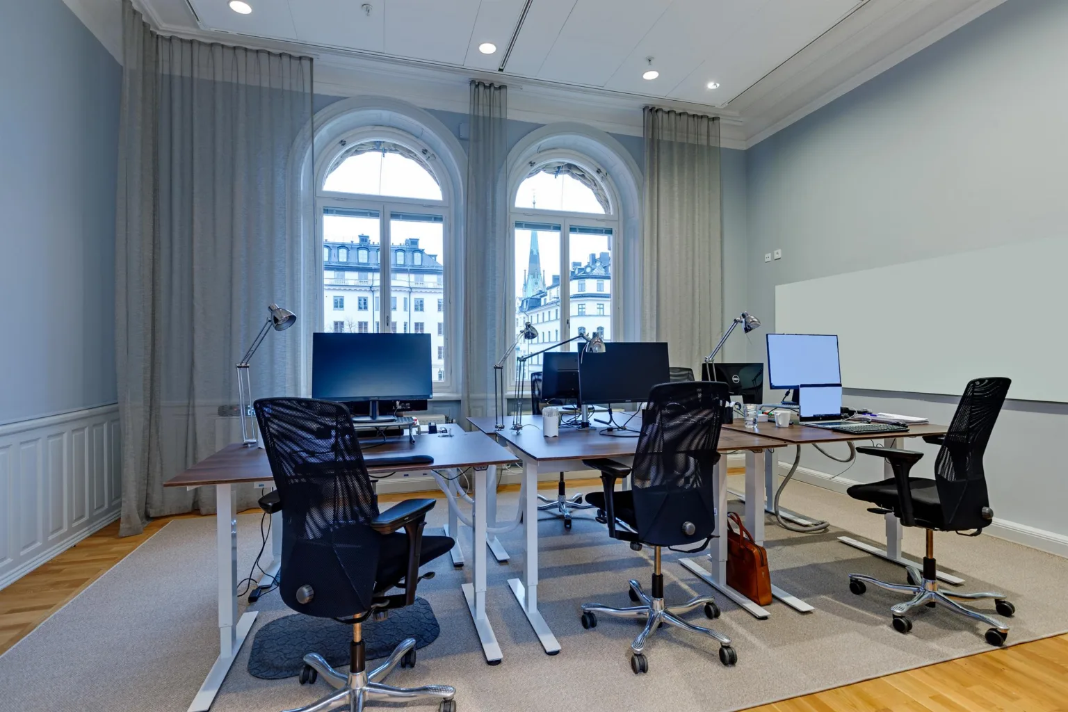 Ett modernt kontor med skrivbord, kontorsstolar och datorskärmar framför välvda fönster som släpper in dagsljus.