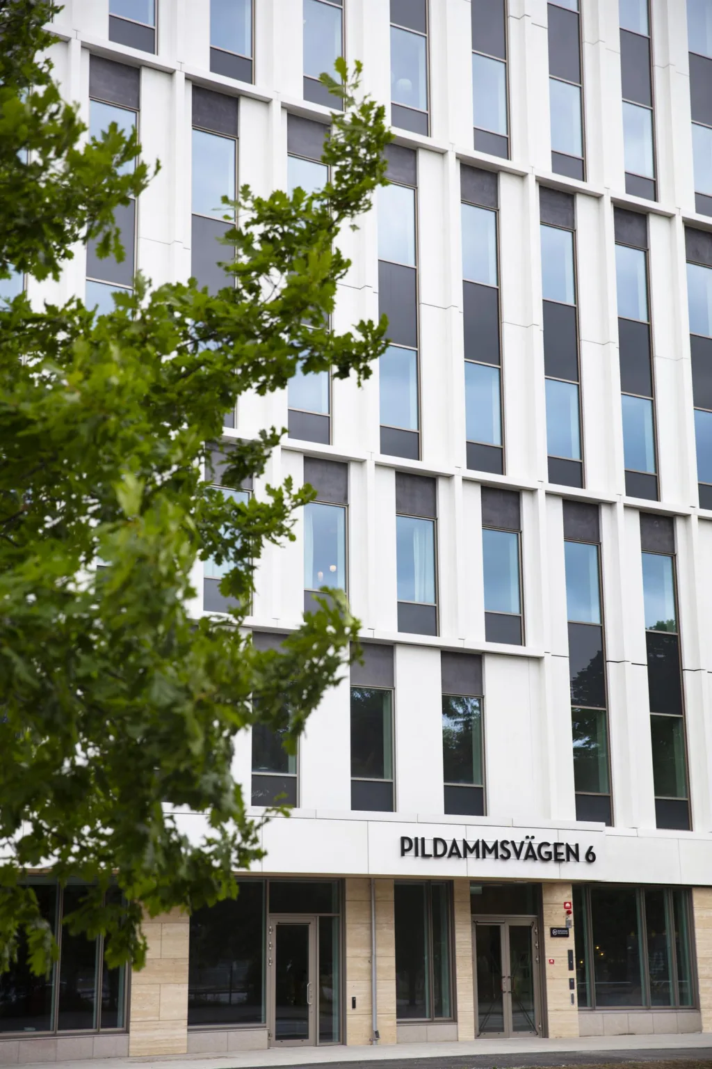 En modern byggnad med vita fasader och stora fönster, texten "Pildammsvägen 4" syns längst ner.