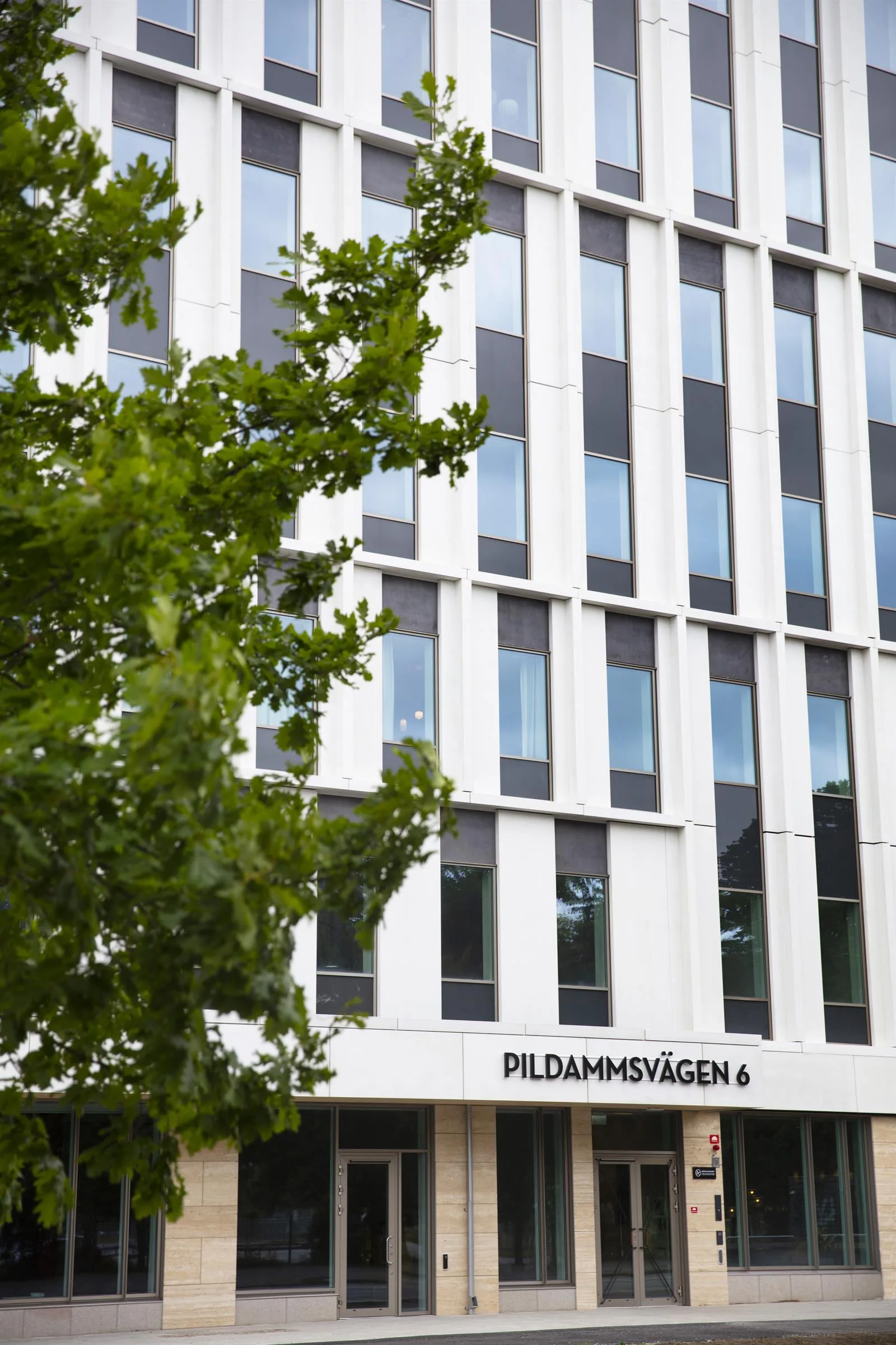 En modern byggnad med vita fasader och stora fönster, texten "Pildammsvägen 4" syns längst ner.