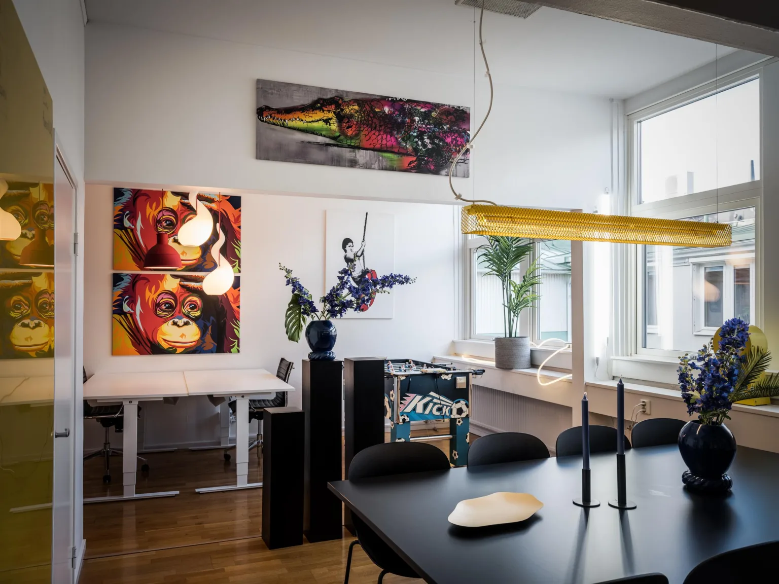 3. Ett rum med ett svart matbord, stolar, färgglada konstverk på väggarna, en blå vas med blommor och ett skrivbord i bakgrunden nära ett fönster.