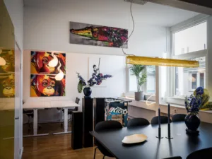 3. Ett rum med ett svart matbord, stolar, färgglada konstverk på väggarna, en blå vas med blommor och ett skrivbord i bakgrunden nära ett fönster.