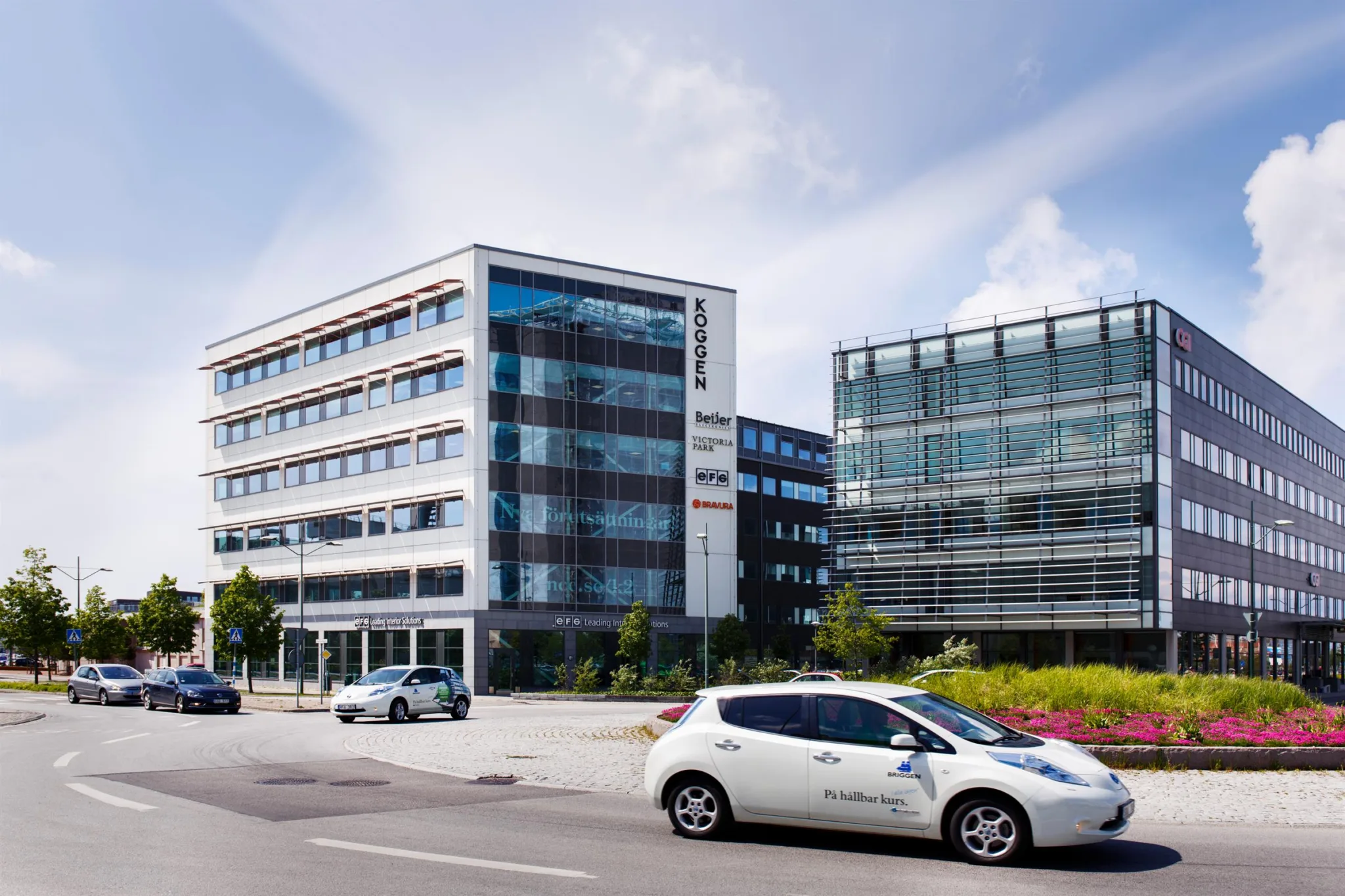 En vit bil står parkerad framför två moderna kontorsbyggnader med glasfasader och en skylt där det står "Innovatum".