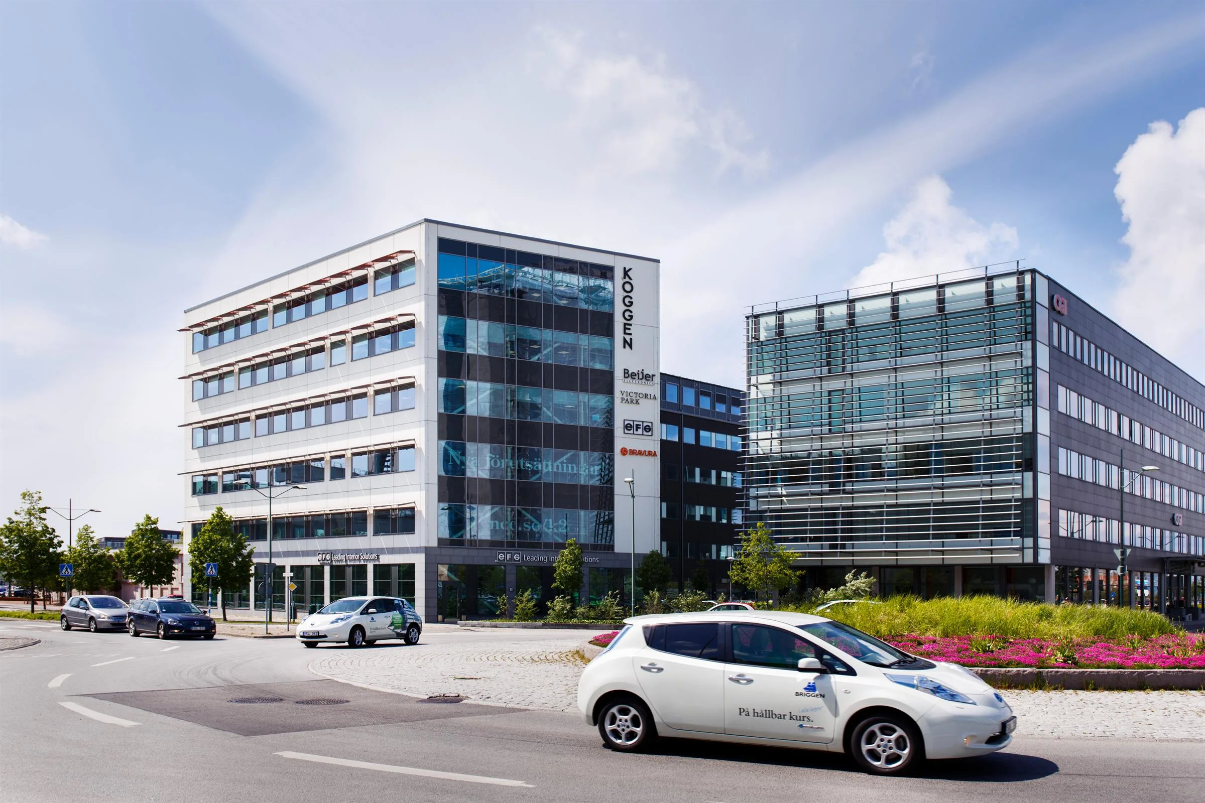 En vit bil står parkerad framför två moderna kontorsbyggnader med glasfasader och en skylt där det står "Innovatum".