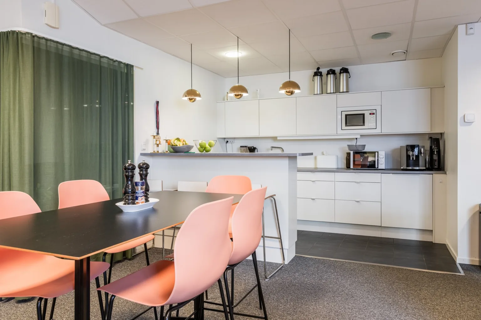 3. Ett kök med vita skåp, rosa stolar vid ett svart bord, hängande lampor och köksutrustning inklusive mikrovågsugn och kaffemaskin.