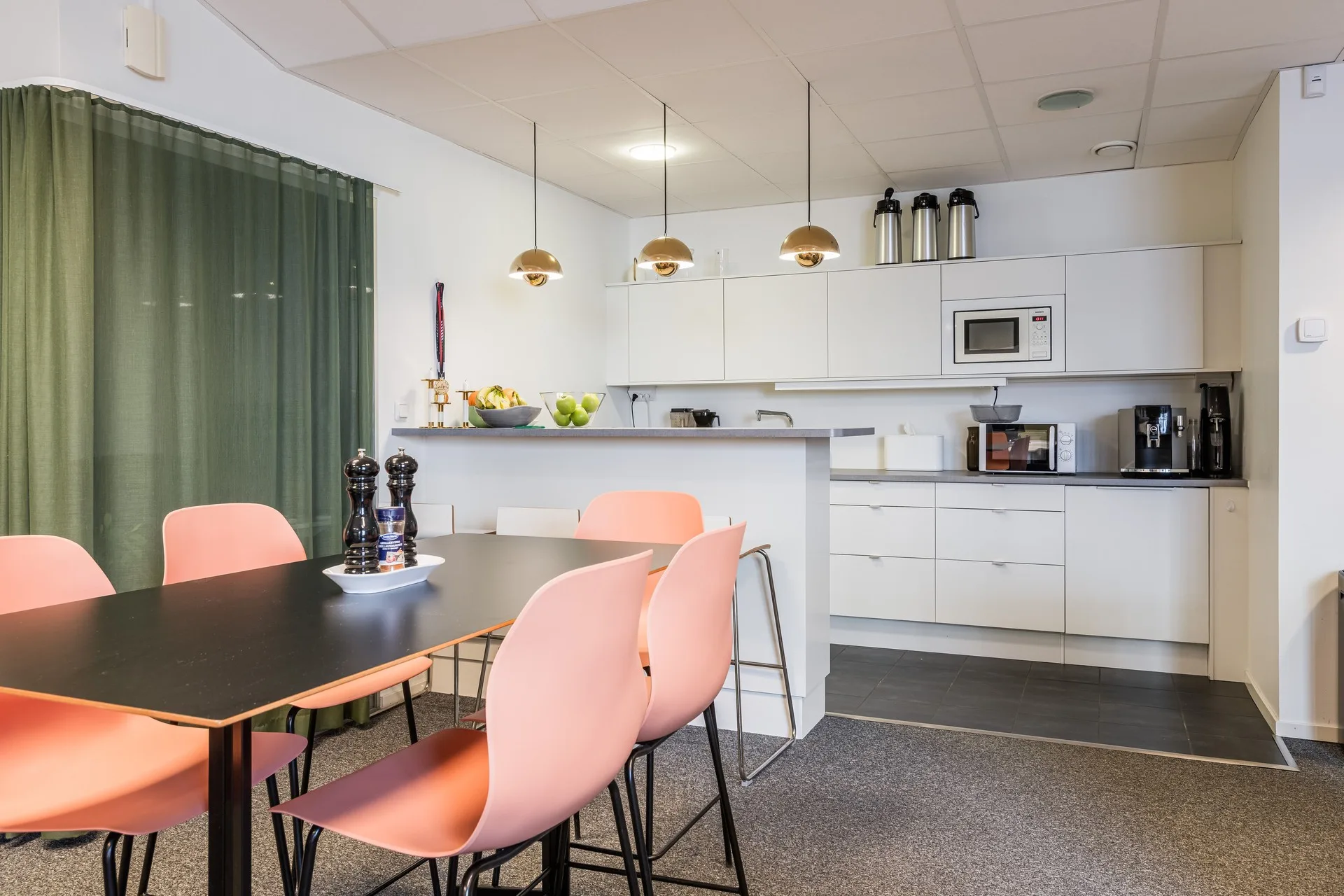 3. Ett kök med vita skåp, rosa stolar vid ett svart bord, hängande lampor och köksutrustning inklusive mikrovågsugn och kaffemaskin.