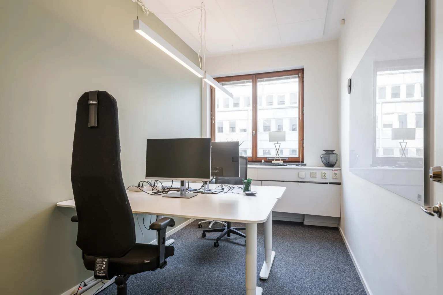 1. Ett kontorsrum med skrivbord, datorskärmar, svart stol och stort fönster som släpper in naturligt ljus.
