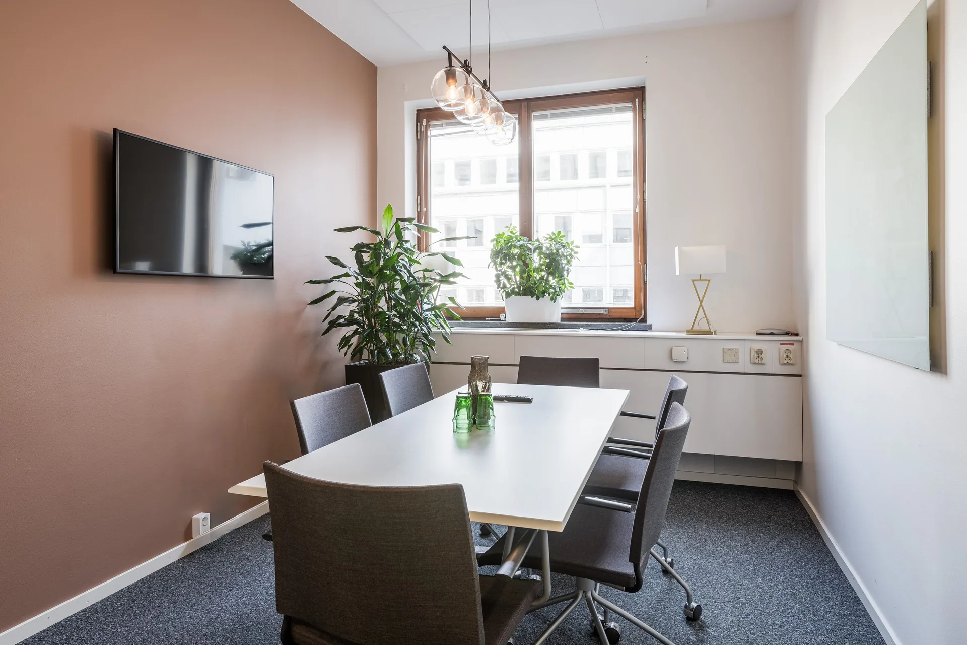 2. Ett mötesrum med ett vitt bord, sex svarta stolar, en väggmonterad TV, växter vid fönstret och en lampa på skänken.