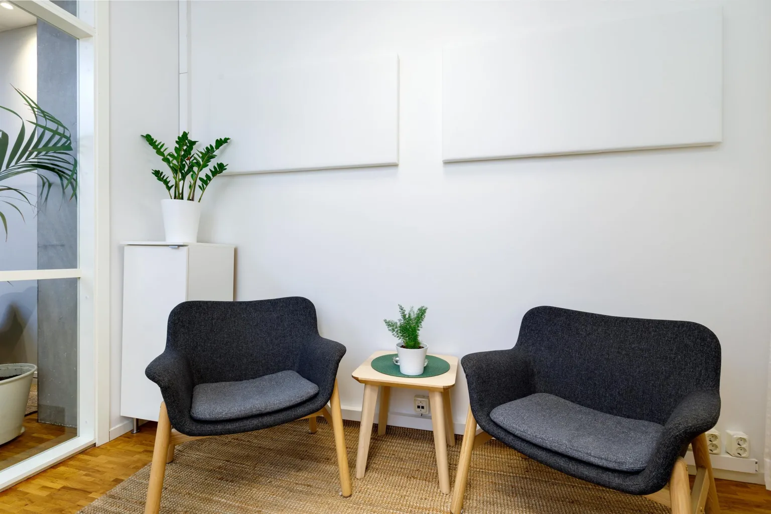 Två grå fåtöljer med ett litet bord emellan, dekorerat med en krukväxt, mot en vit vägg.