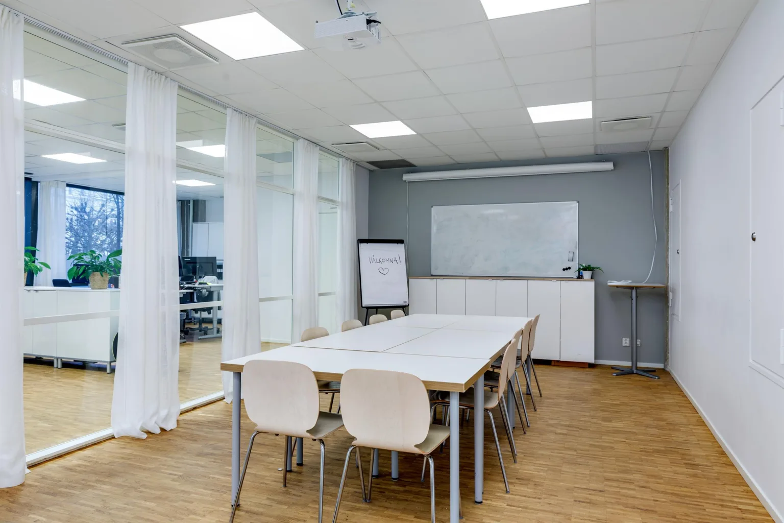 Ett konferensrum med ett vitt bord, stolar, glasväggar, en whiteboard och ett blädderblock.
