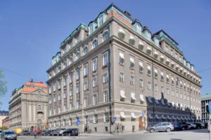 Hörnvy av en stor byggnad med flera våningar och fönster, omgiven av bilar och trottoarer.