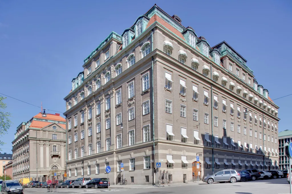 Stor klassisk byggnad med flera våningar, grå fasad, och grönt tak, omgiven av parkerade bilar.