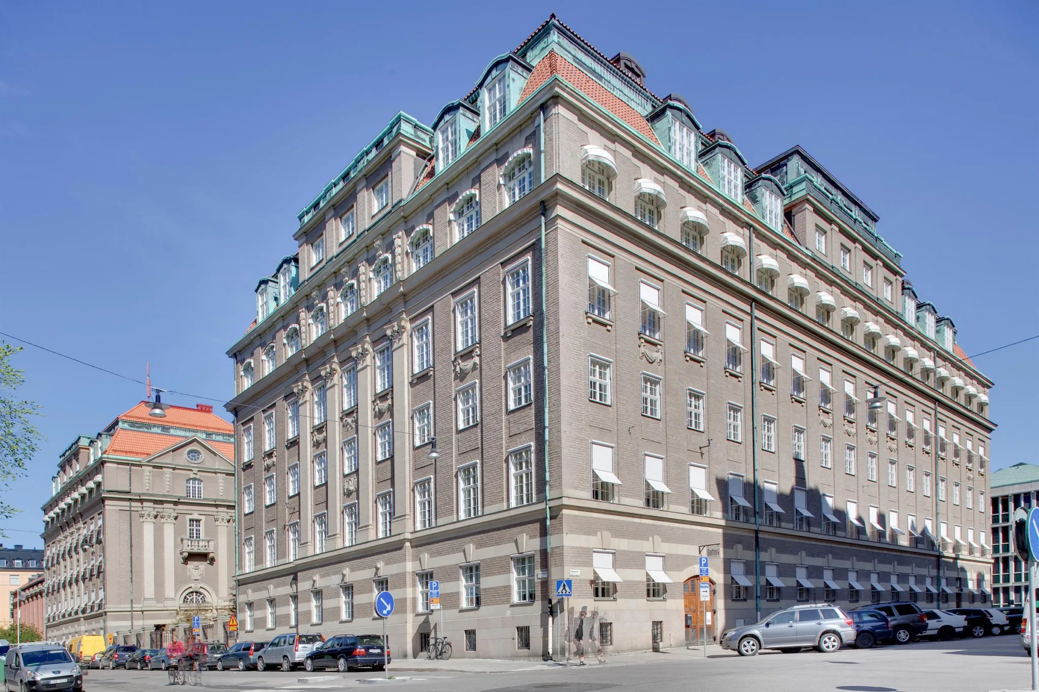 Stor klassisk byggnad med flera våningar, grå fasad, och grönt tak, omgiven av parkerade bilar.