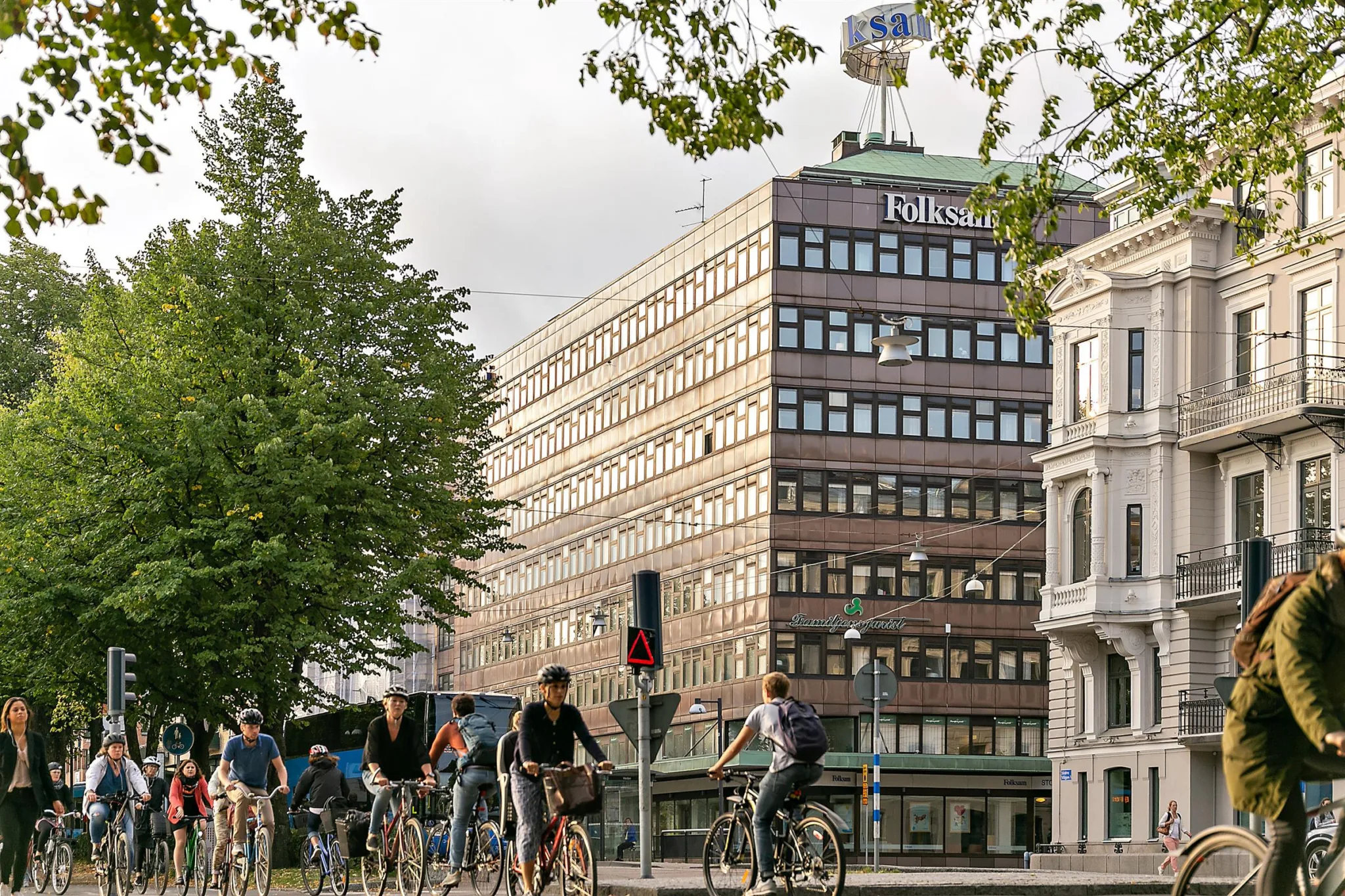Cyklande människor på en stadsgata framför en större brun byggnad med skyltar och grönska.