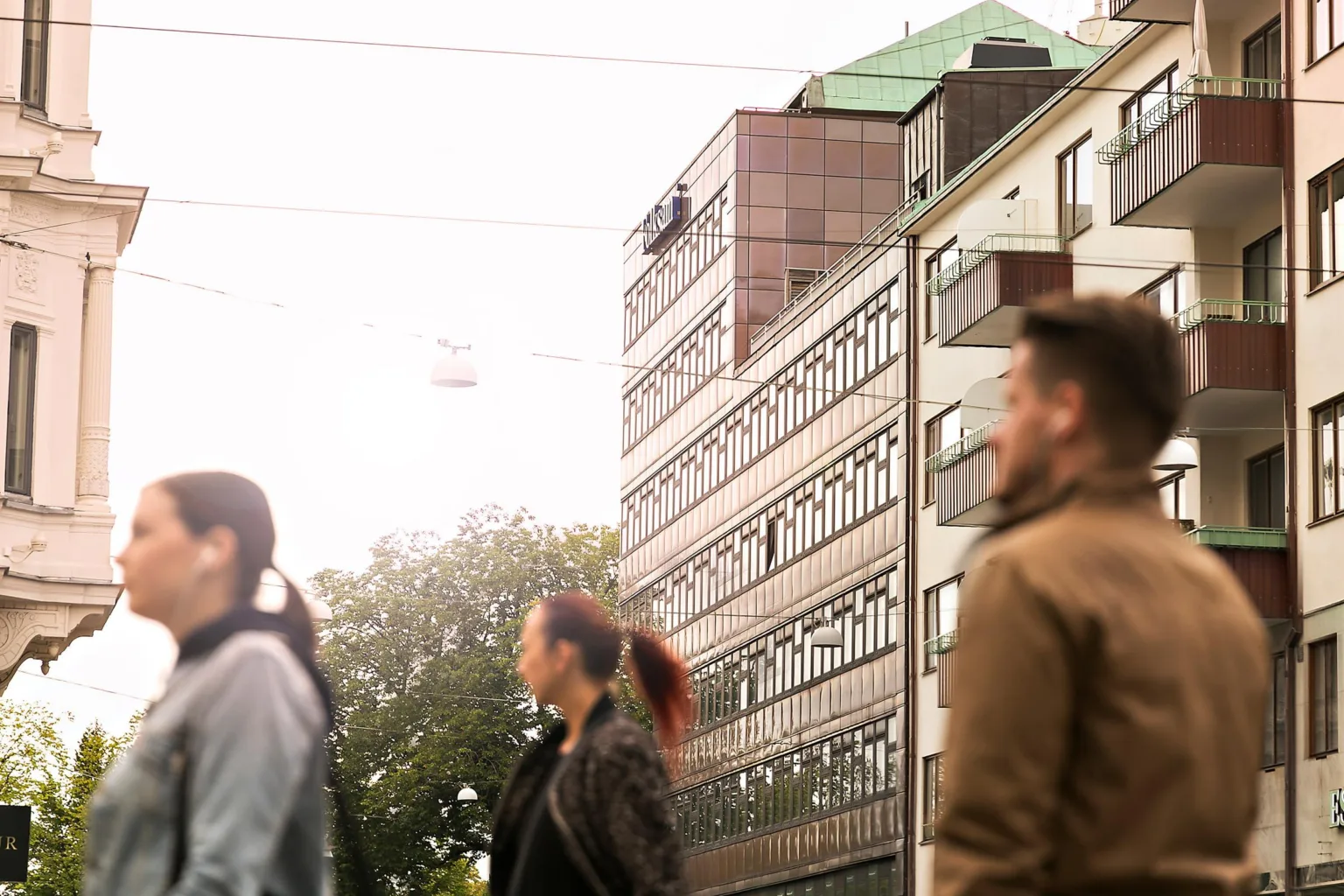 Folk som går på en stadsgata, med en modern byggnad i bakgrunden under en ljus himmel.