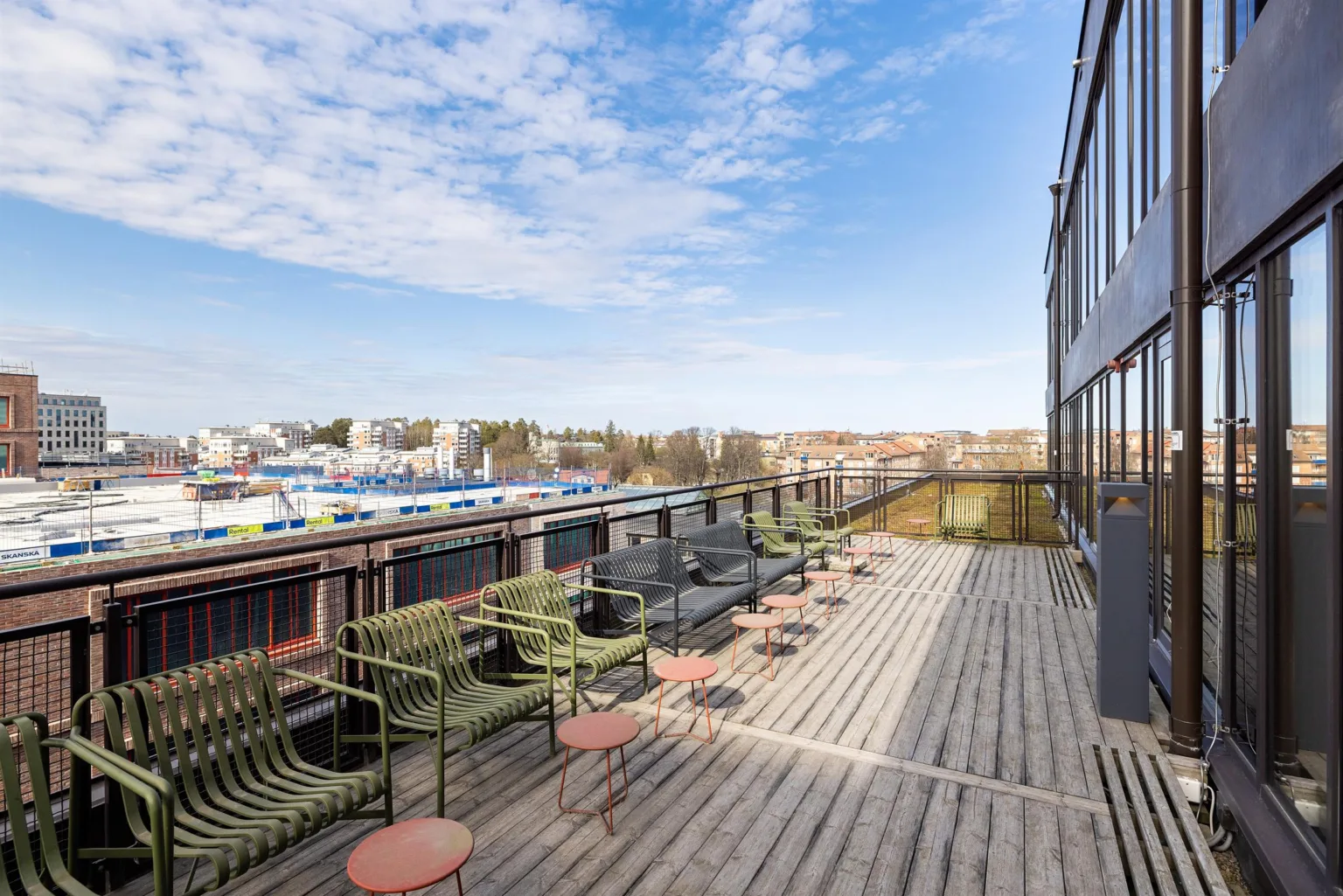 1. En utomhus terrass med trädäck, gröna stolar, små runda bord och utsikt över ett stadsområde under en blå himmel.