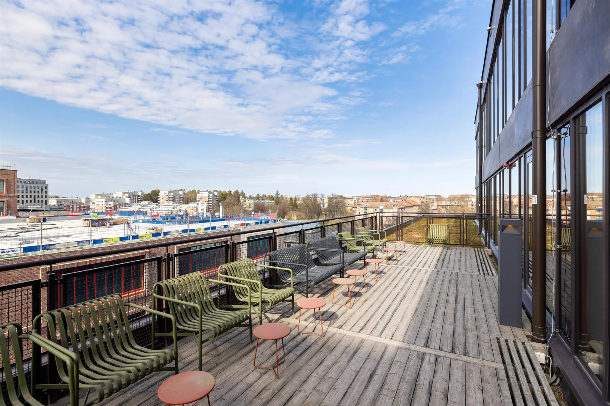 1. En utomhus terrass med trädäck, gröna stolar, små runda bord och utsikt över ett stadsområde under en blå himmel.