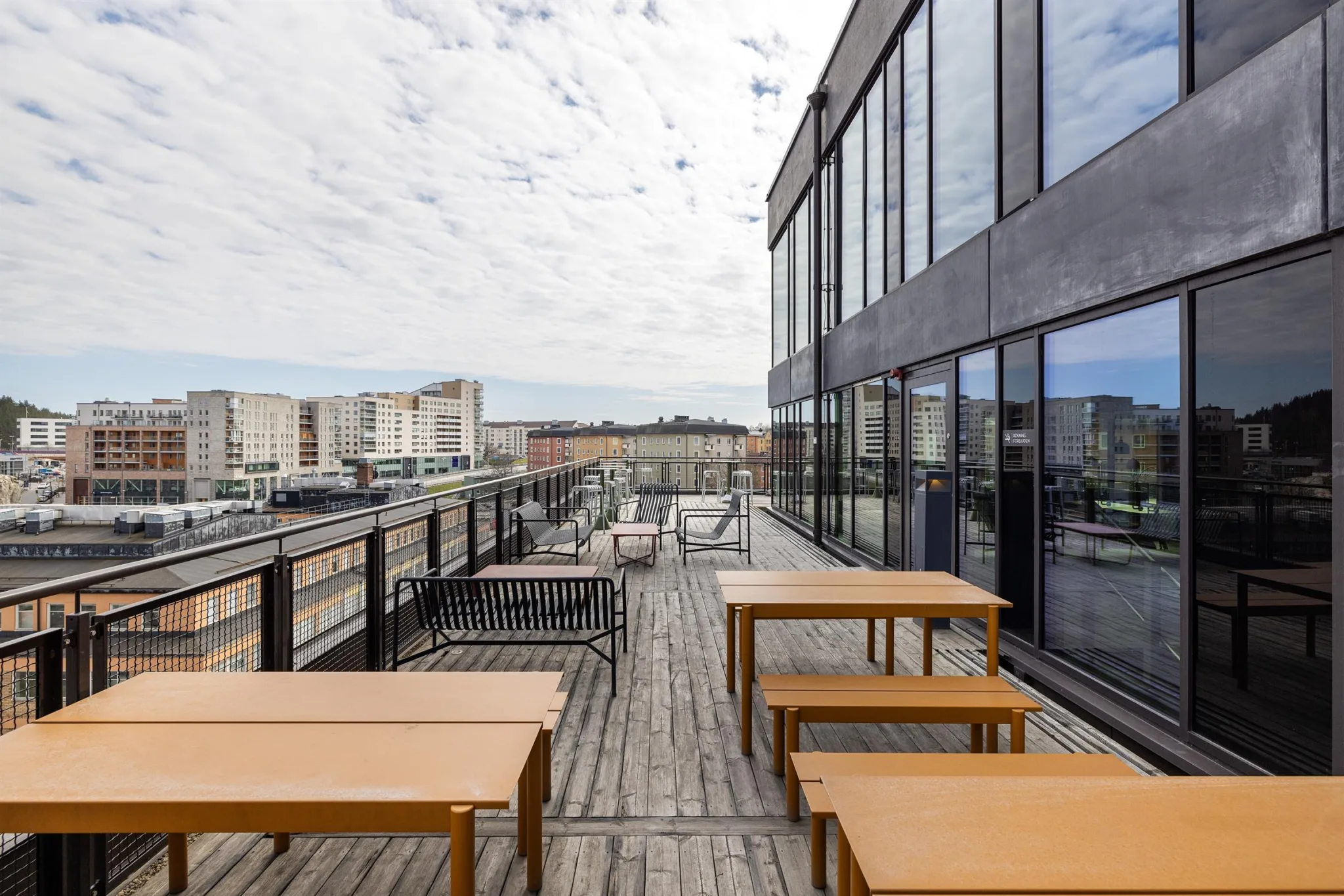 En takterrass med träbord och bänkar, omgiven av glasräcken och utsikt över staden under en molnig himmel.