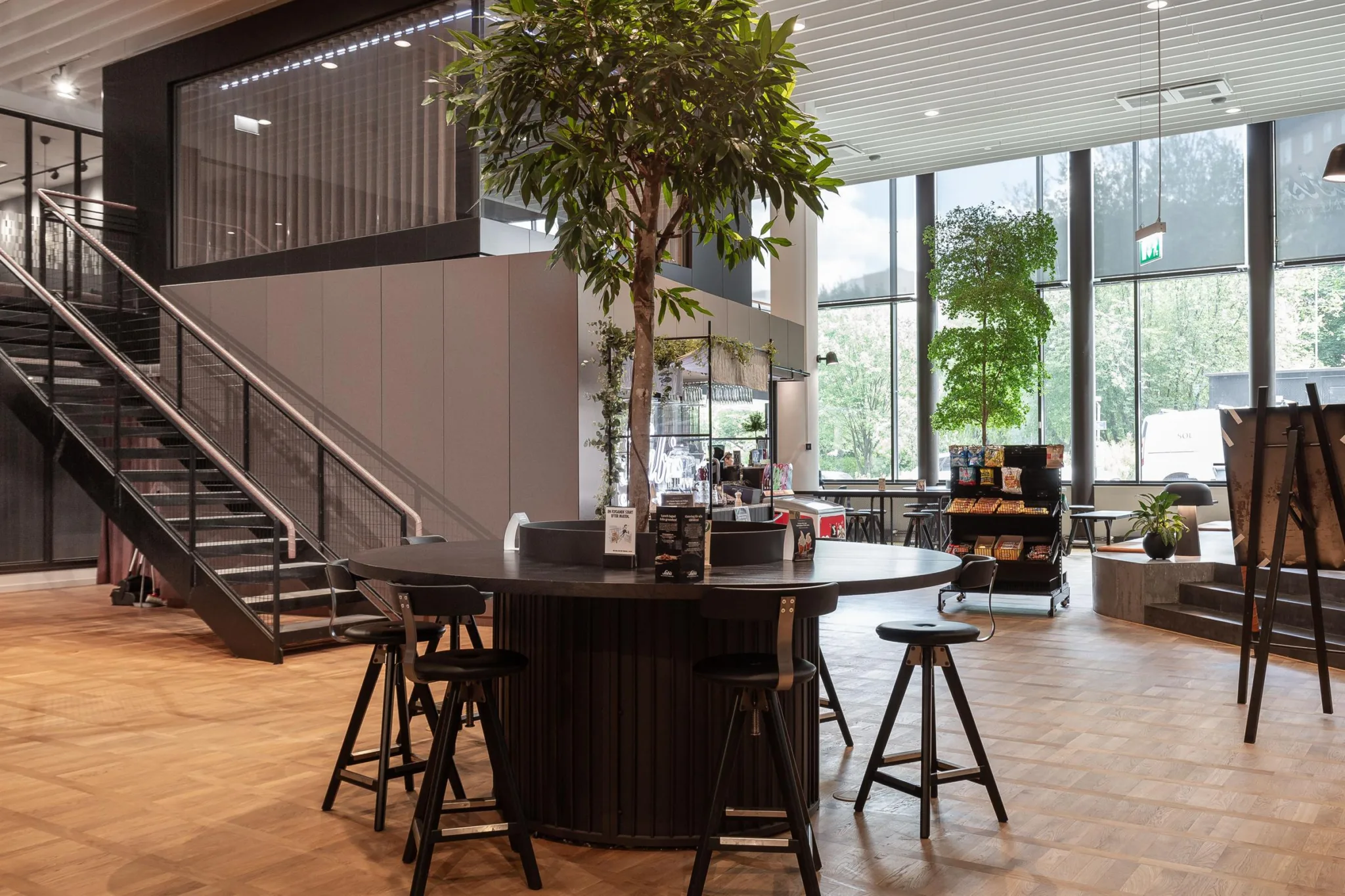 5. En modern lounge med ett runt bord, barstolar och ett träd i mitten, omgiven av glasväggar och stora fönster.