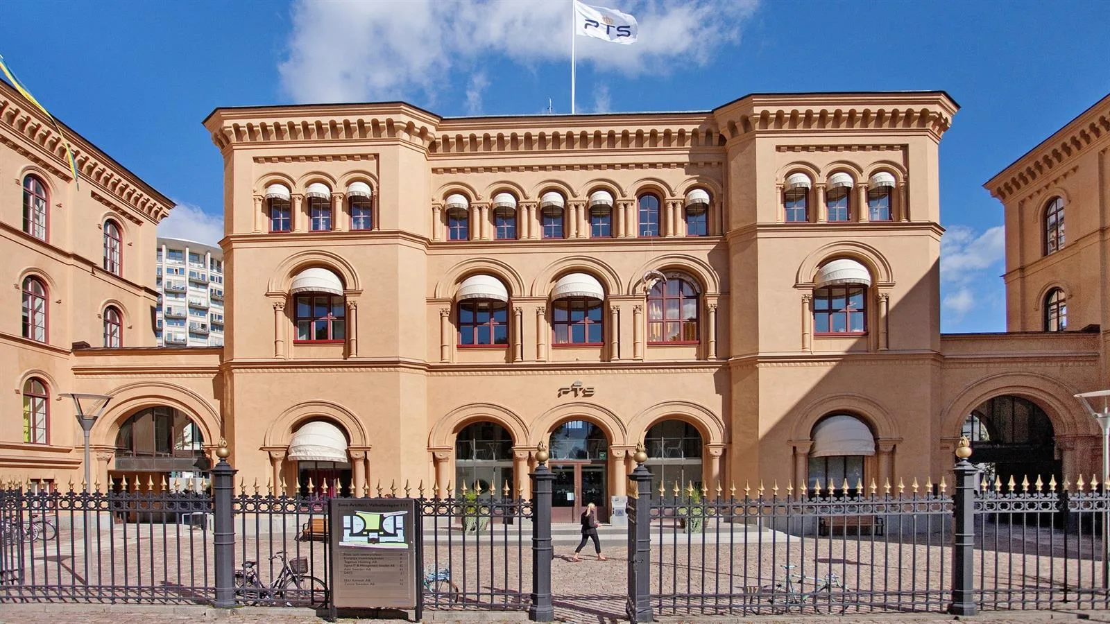 En historisk byggnad med beige fasad och välvda fönster. En flagga vajar på taket och en grind syns i förgrunden.
