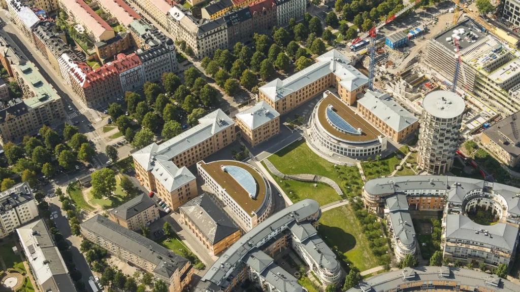 Flygfoto över ett campusområde med flera byggnader, grönområden och träd. Byggnaderna har böjda tak och är omgivna av stadsmiljö.