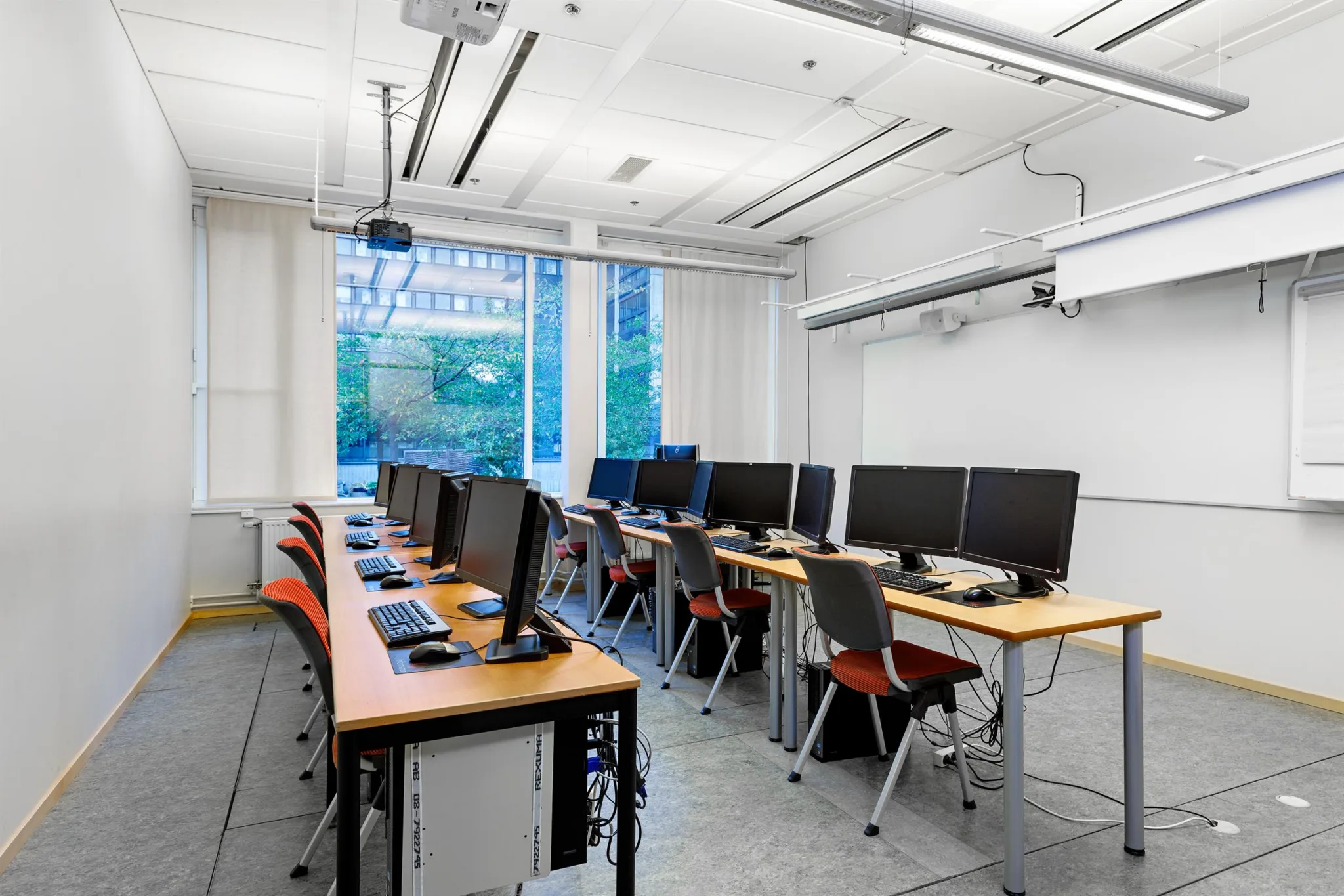 3. Ett ljust datorklassrum med flera skrivbord och datorskärmar, samt stora fönster i bakgrunden.