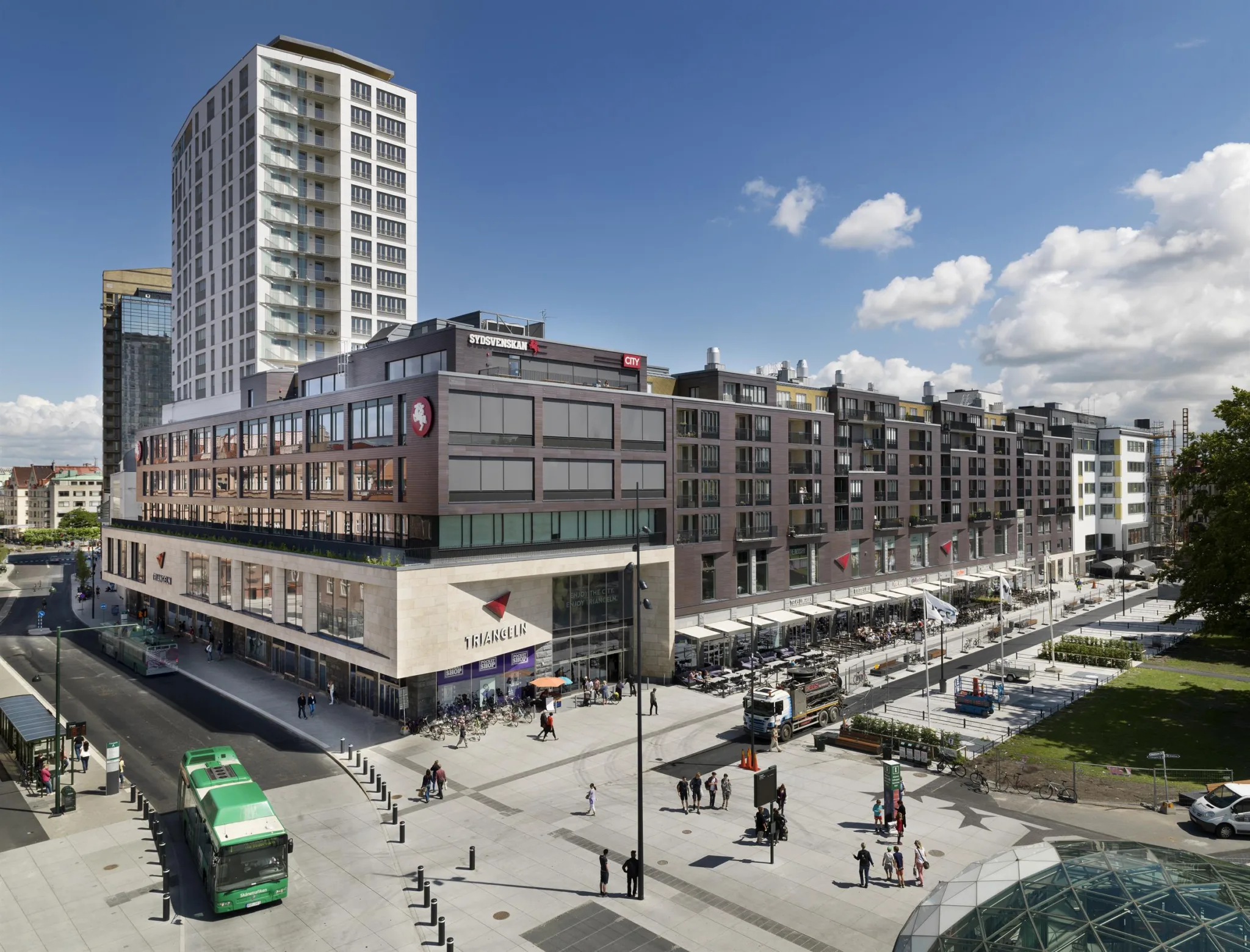 2. Ett stadstorg med höga byggnader, butiker och spårvagn i rörelse under en blå himmel.