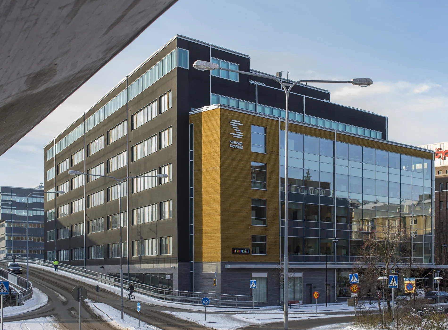 En modern byggnad med stora glaspartier och svart fasad står vid en snötäckt gata.