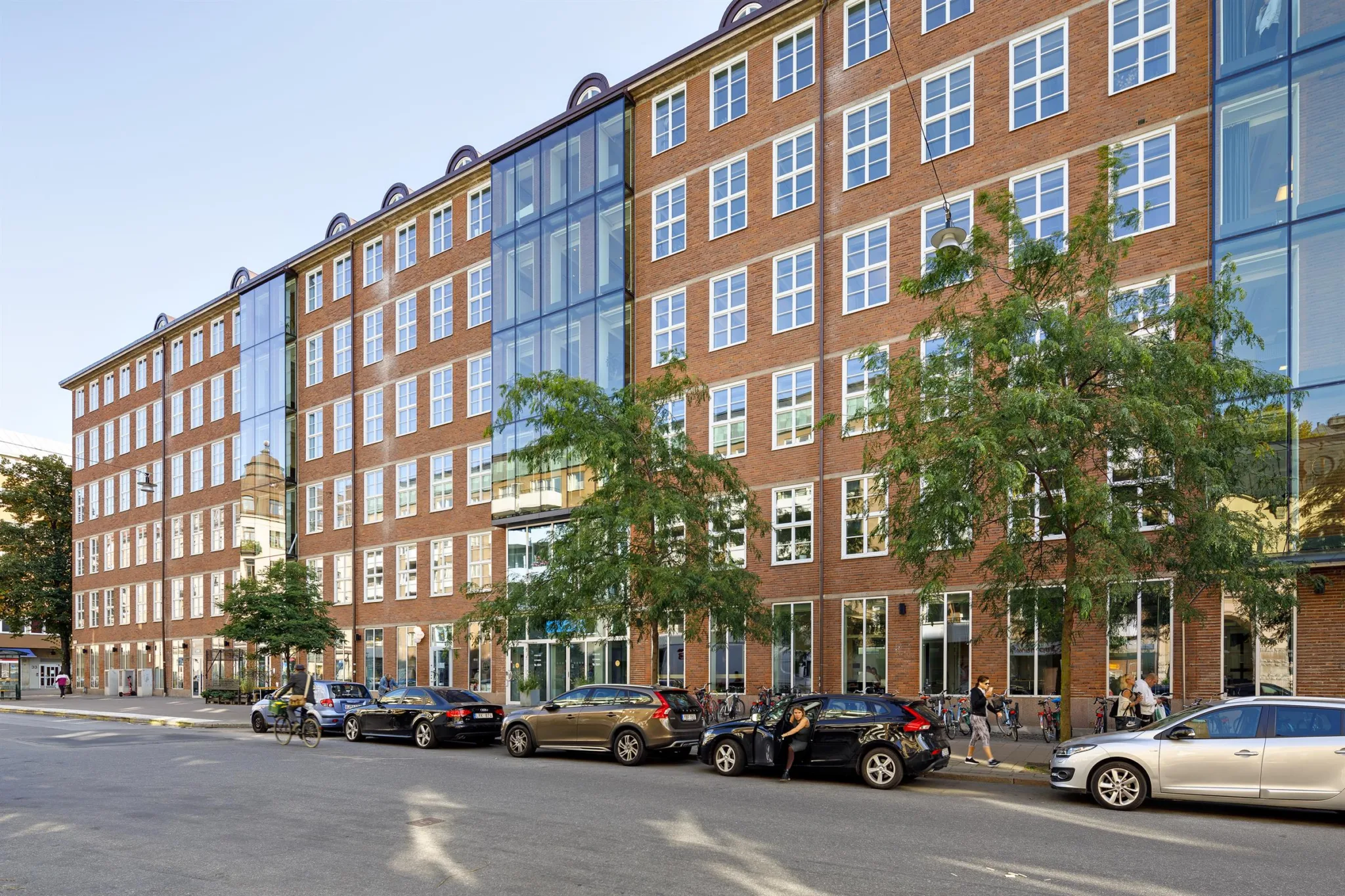 En tegelbyggnad med stora glaspartier, träd och parkerade bilar längs gatan.