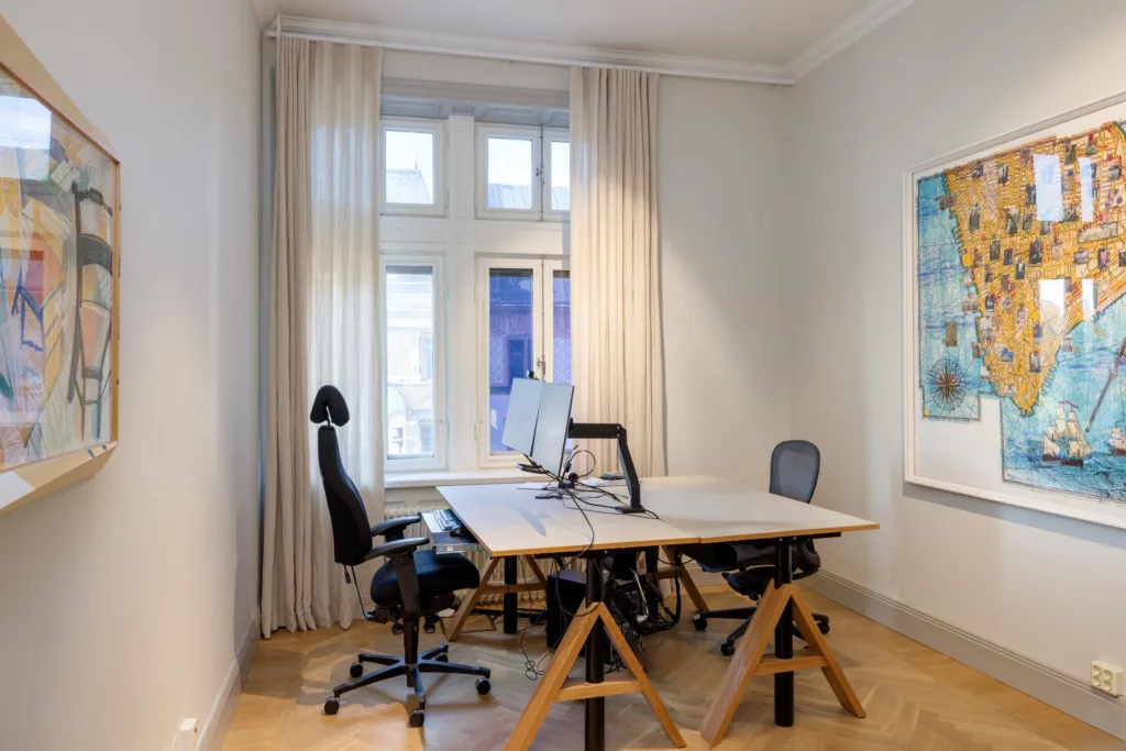 Ett ljust kontorsrum med två skrivbord, ergonomiska stolar och datorer, framför ett stort fönster med vita gardiner.