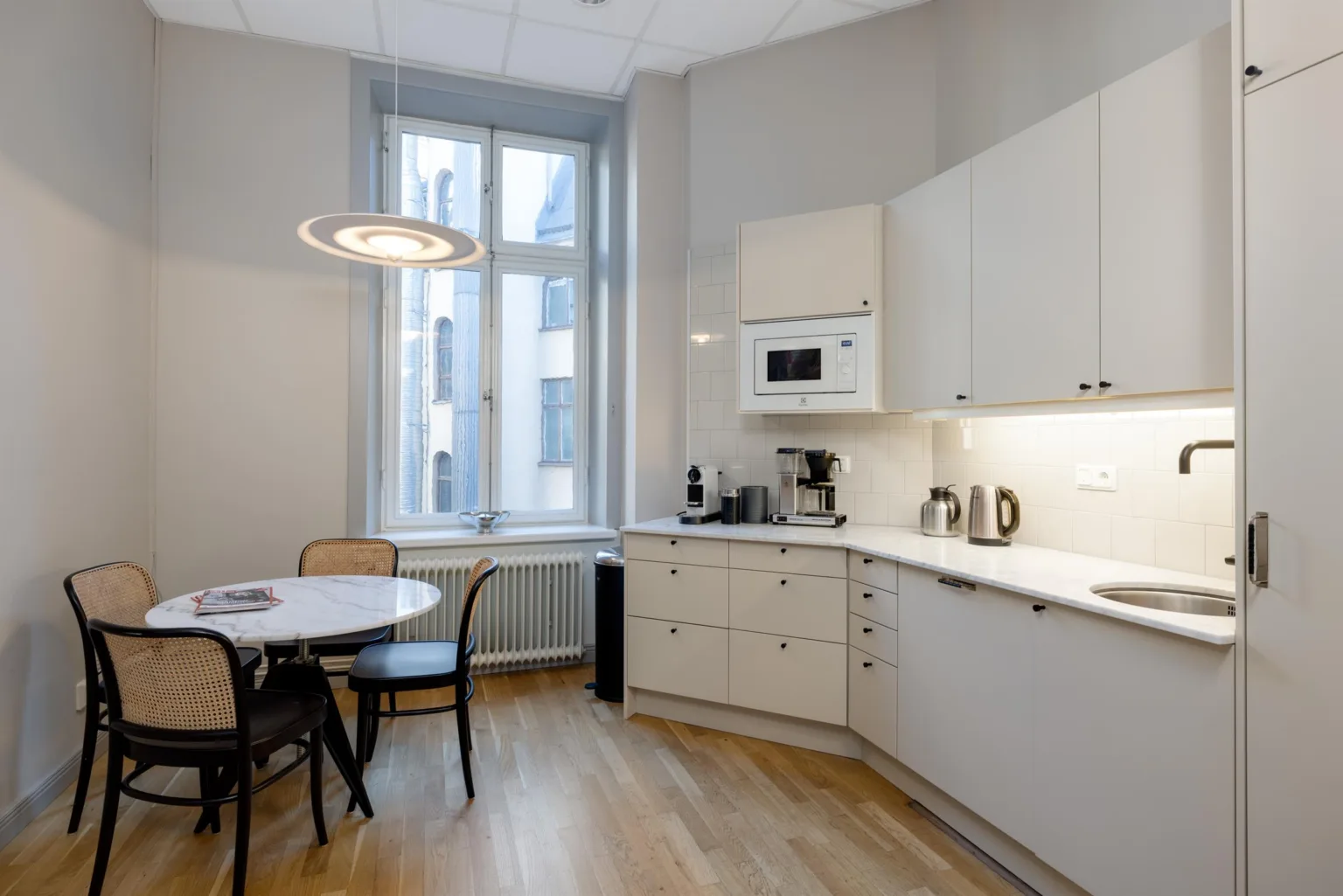 Ett kök med vita skåp, en mikro, vattenkokare, kaffemaskin samt ett matbord med tre stolar vid ett fönster.