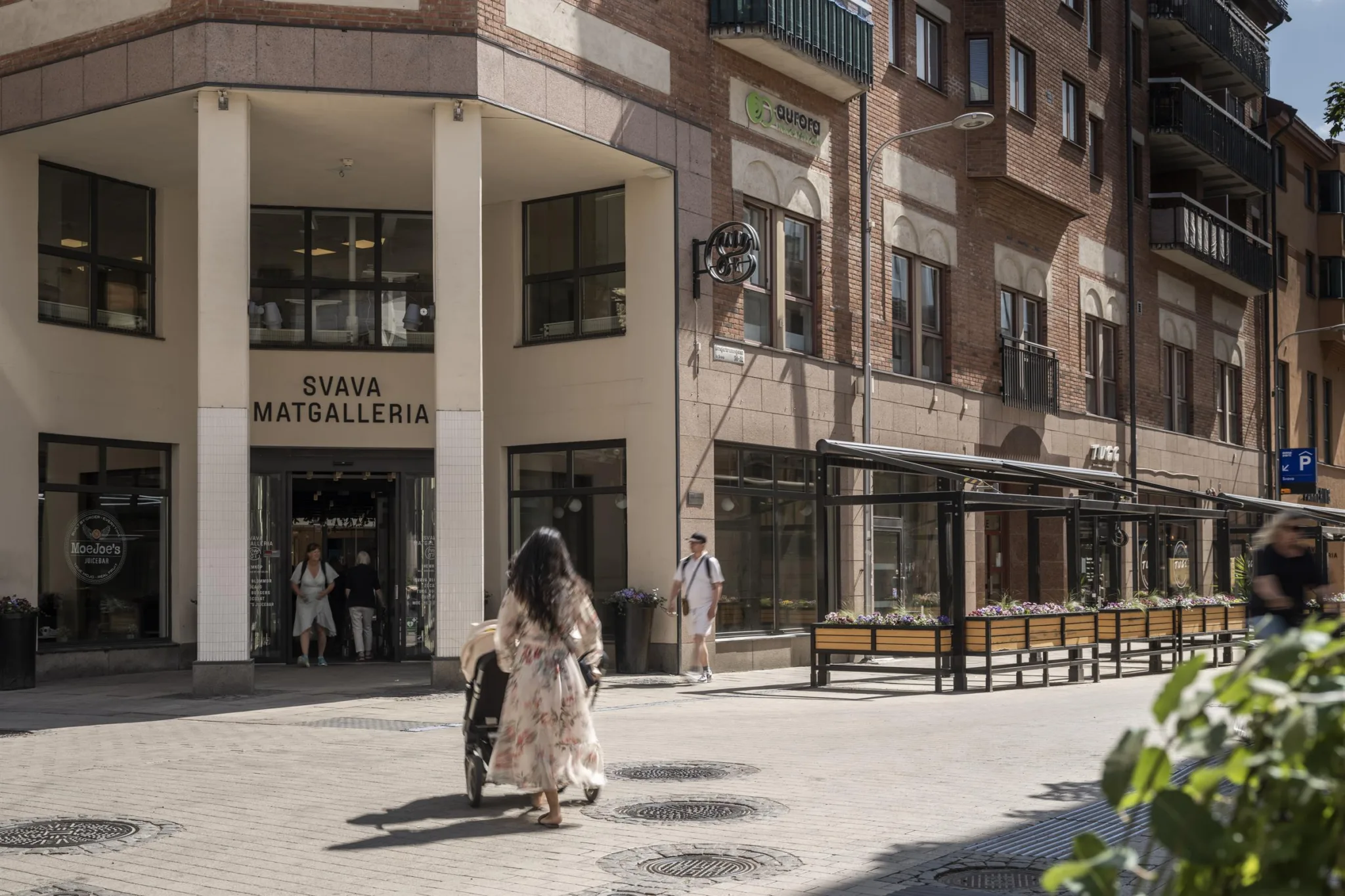 En kvinna med långt hår och ljus klänning går mot ingången till "TÄBY MATGALLERI" på en solig dag.