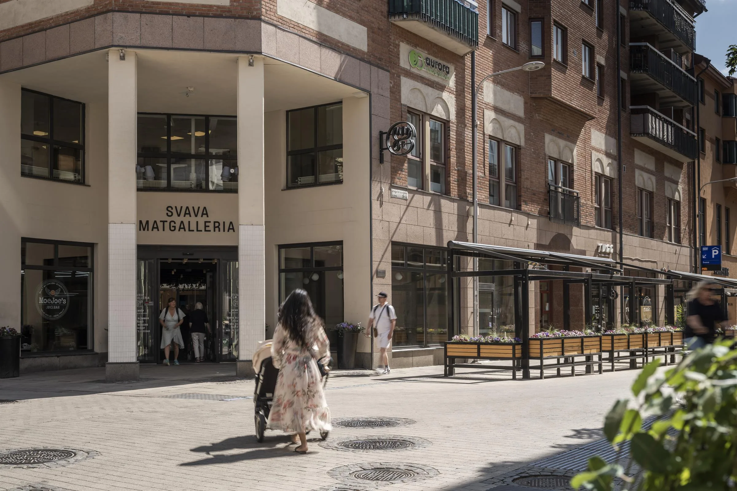 En kvinna med långt hår och ljus klänning går mot ingången till "TÄBY MATGALLERI" på en solig dag.