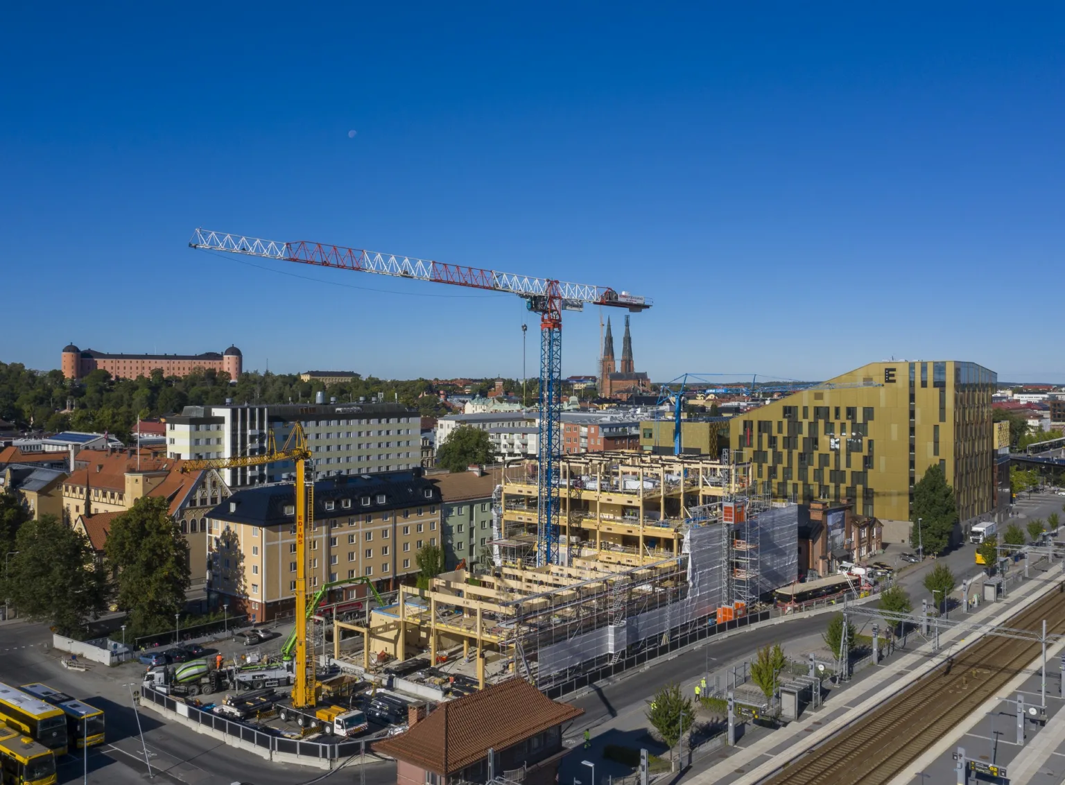 En byggarbetsplats med två höga kranar, omgivna av stadsmiljö och blå himmel i bakgrunden.